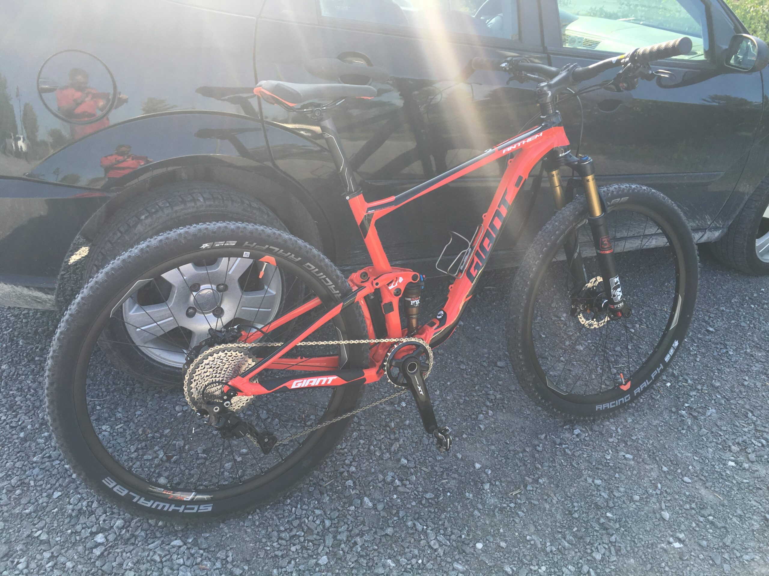Giant Anthem: A bright orange mountain bike leaning against a black car, showcasing its front and back wheels. The bike features a lightweight frame, prominent suspension, and various gear components. The background includes gravel and the reflection of trees and a person in the car's surface.