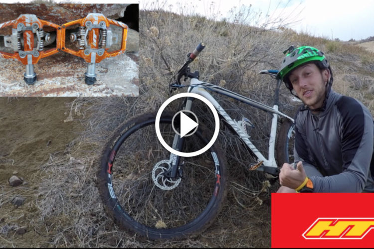 A cyclist wearing a helmet and a long-sleeve shirt sits next to a mountain bike on a rocky terrain. An inset image displays a pair of orange bike pedals. A play button overlay indicates this is a video thumbnail, and a logo for "HT" is visible at the bottom right corner.