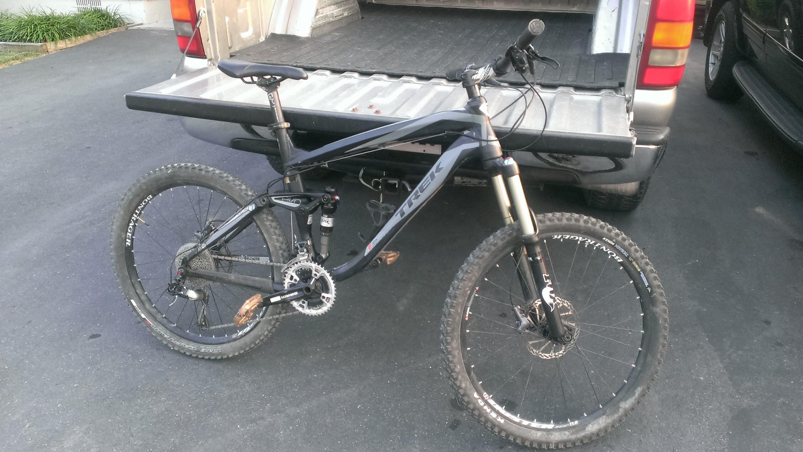 Trek Remedy 7: A black mountain bike is leaning against the open tailgate of a pickup truck. The bike features thick, knobby tires and a suspension system, indicating it's designed for off-road use. The scene is set on a paved surface, with some greenery visible in the background.