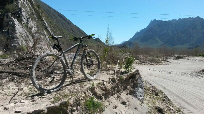 Scott Scale 950: A mountain bike leaning against a rocky ledge on a sandy path, surrounded by sparse vegetation and mountains in the background under a clear blue sky.