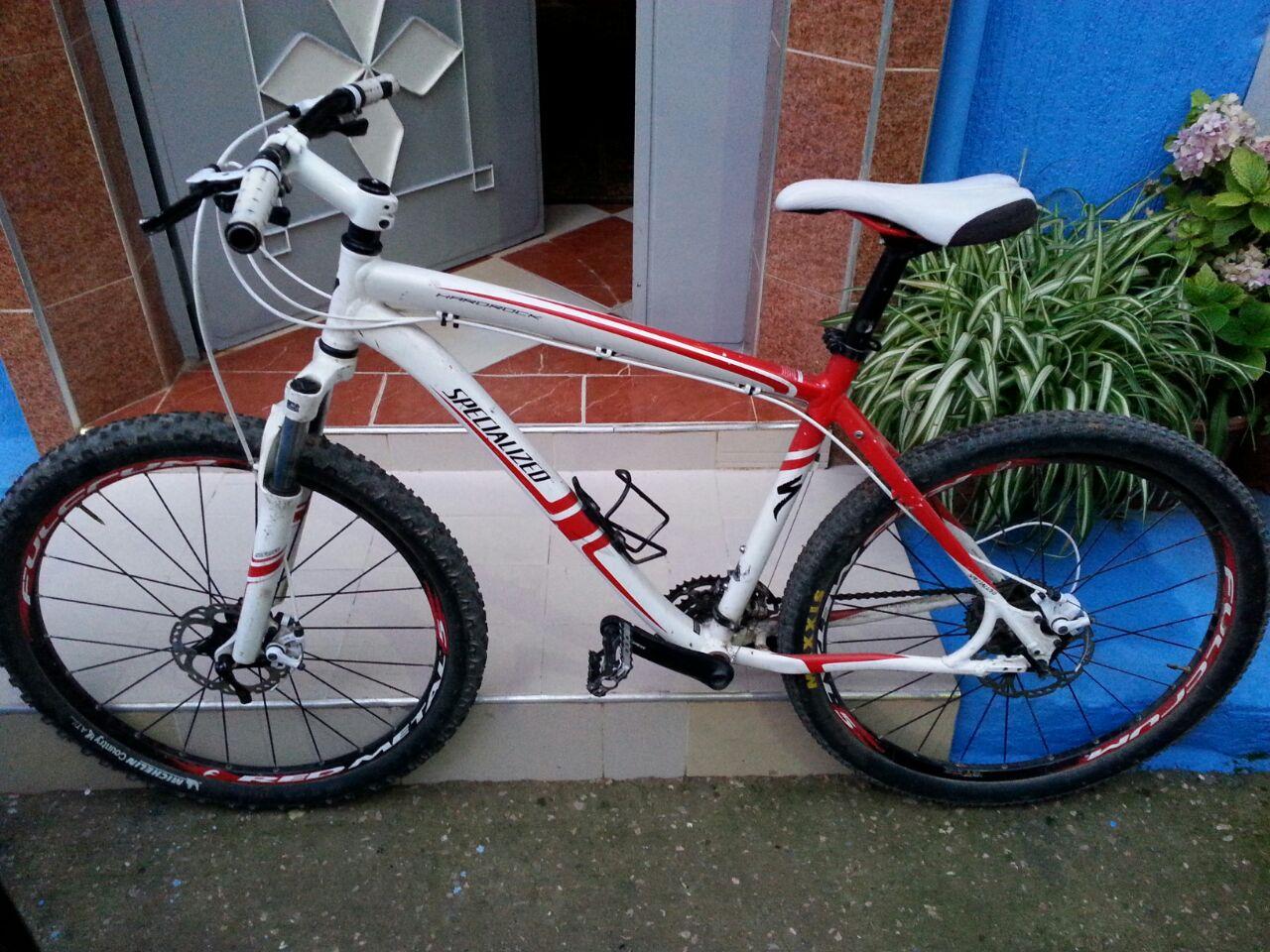 Specialized hardrock: Mountain bike leaning against a wall, featuring a white and red frame, thick black tires, and visible gears and brakes. The background includes a blue wall and some greenery.