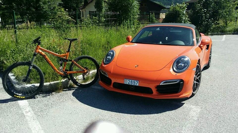 Kona Process 134 DL: An orange Porsche sports car parked next to an orange mountain bike in a sunny outdoor setting with green grass and trees in the background.