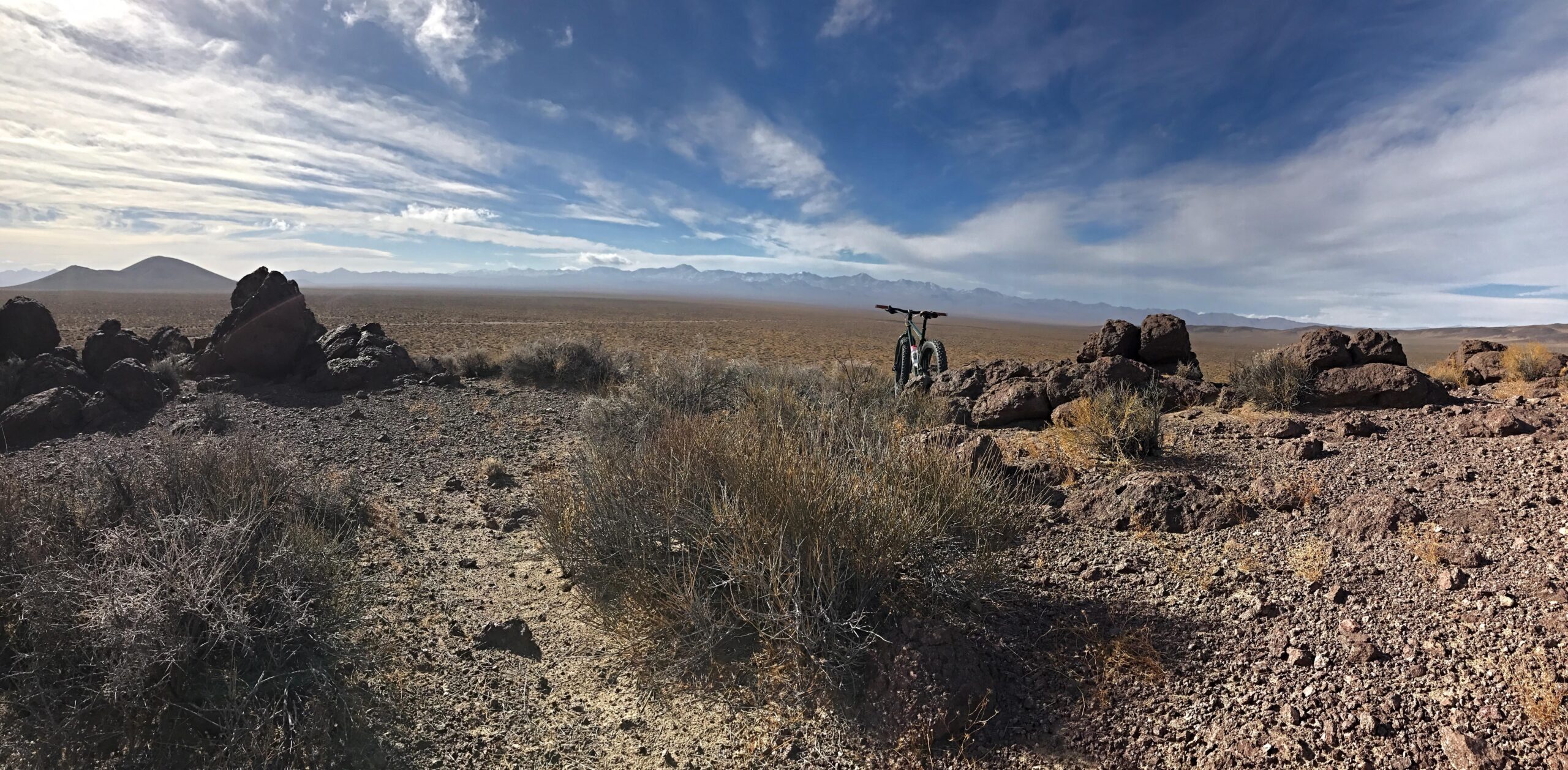 Nashbar Big Ol Fat Bike: A panoramic view of a rugged, arid landscape featuring rocky formations and sparse vegetation, with a distant mountain range under a partly cloudy sky. A bicycle is positioned on the right side of the image, suggesting an outdoor adventure in a remote area.