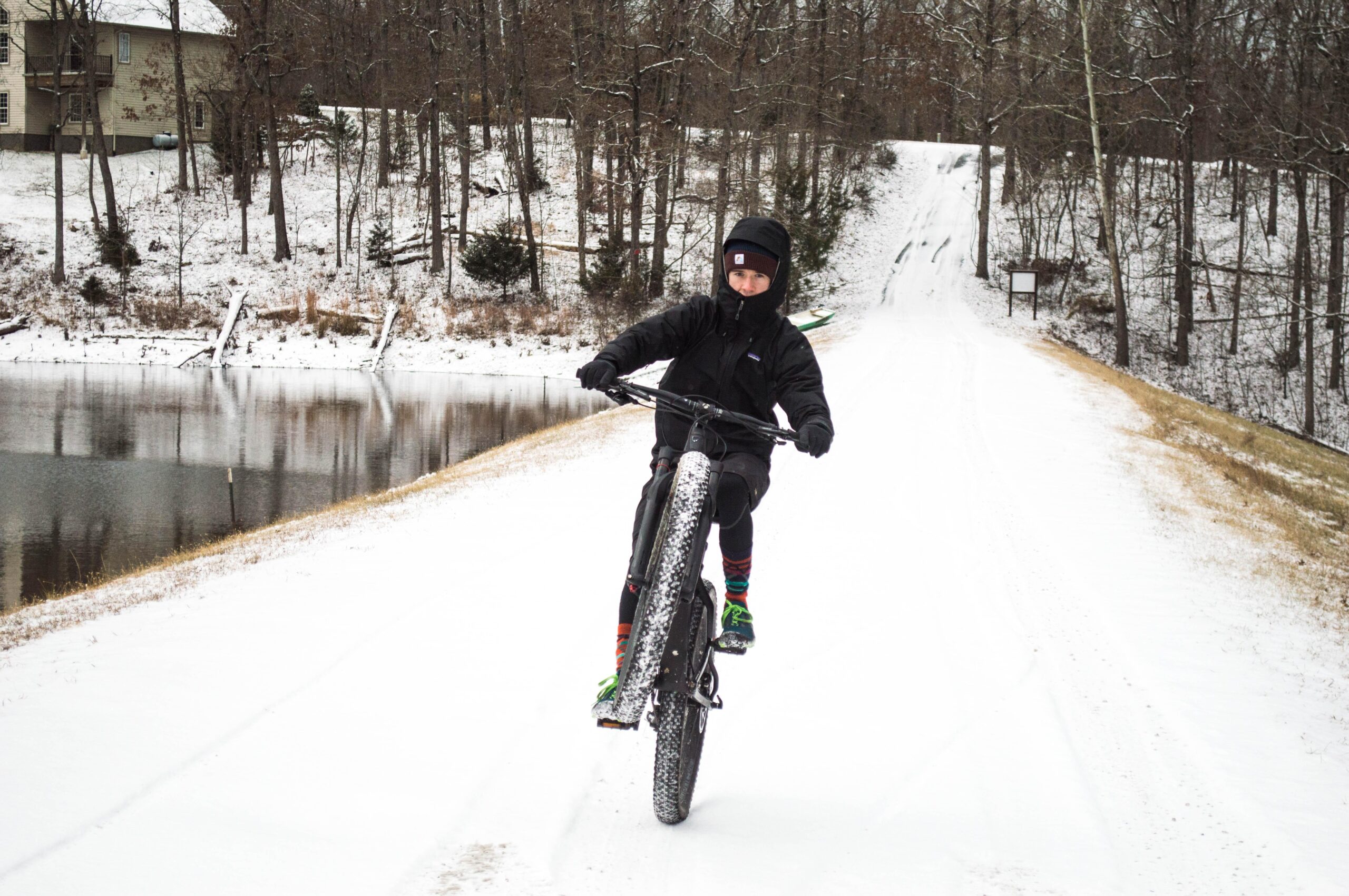 Trek Stache 9: A person wearing a black jacket and beanie rides a fat-tire bike on a snow-covered path next to a pond, performing a wheelie. Snow-covered trees line the background, and a house is visible on the left side of the image.