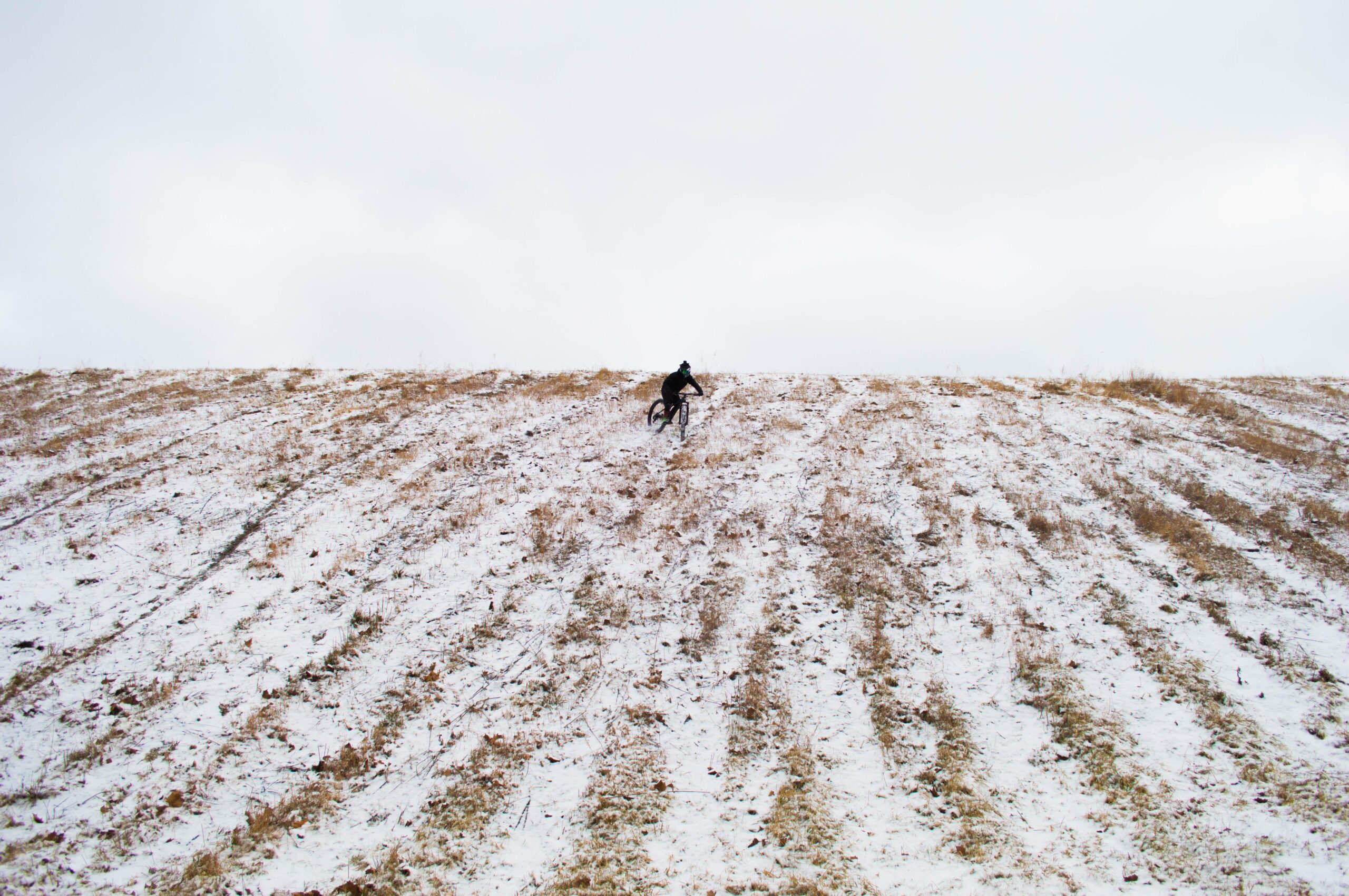 Trek Stache 9: A cyclist rides down a snowy, sloped field with sparse vegetation under a cloudy sky.