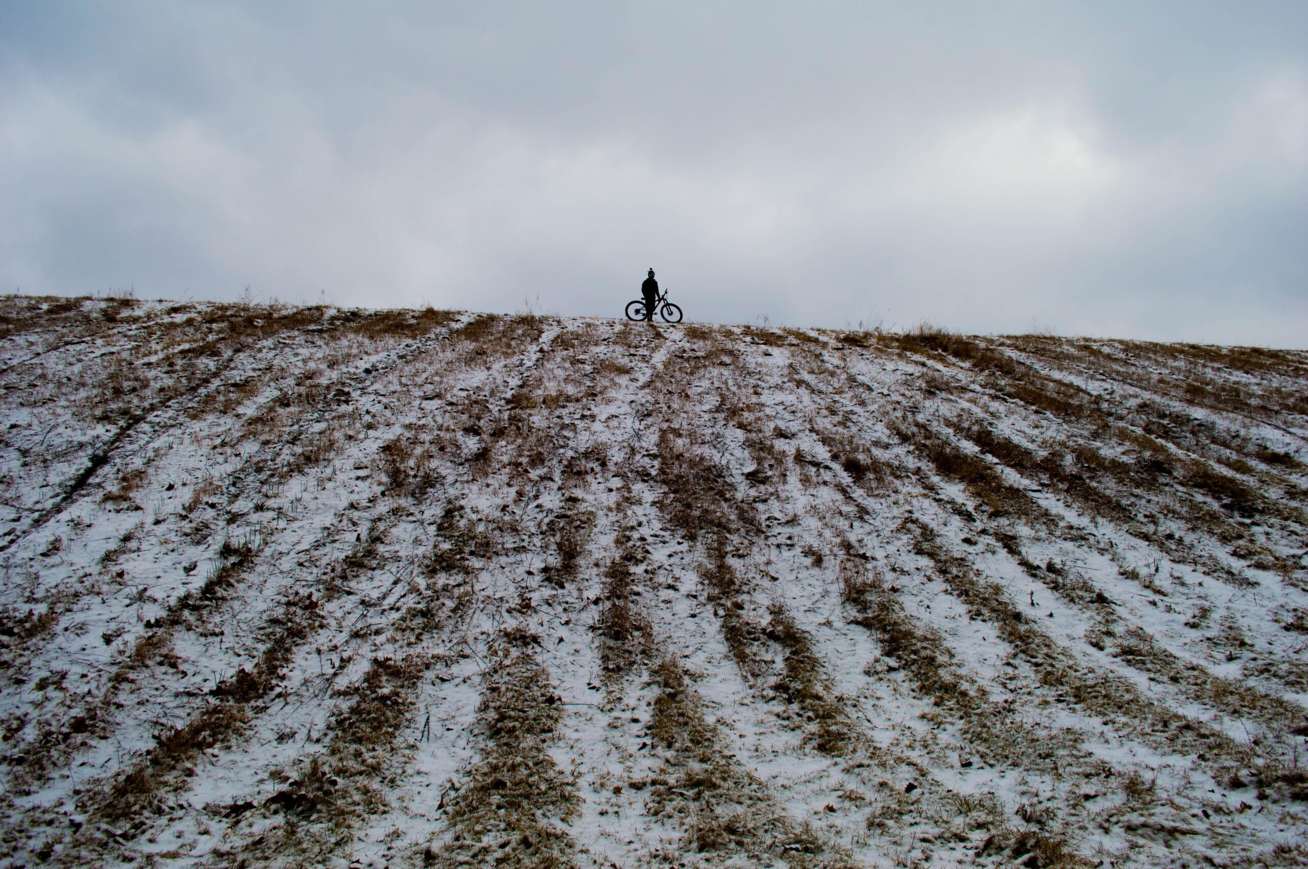 Trek Stache 9: A silhouette of a person riding a bike on a snowy hillside, with a cloudy sky in the background. The hill shows visible tracks and patches of snow amidst the grass.