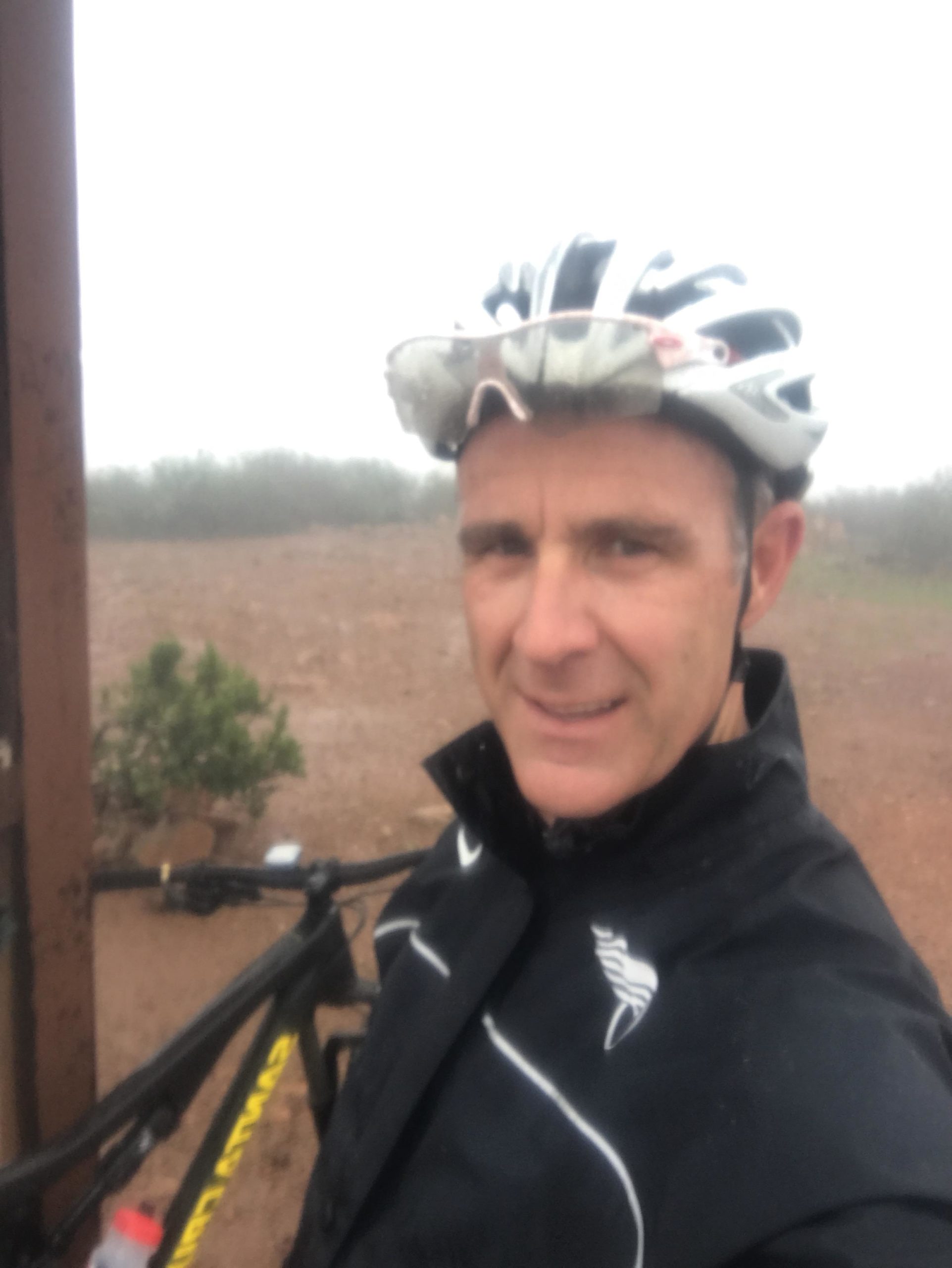 A person wearing a cycling helmet and a dark jacket with white accents stands next to a mountain bike. The background is foggy and has a blurred landscape with sparse vegetation, suggesting rainy or misty weather. The individual is smiling, posing for a selfie. Discovery Lake mountain bike trail.