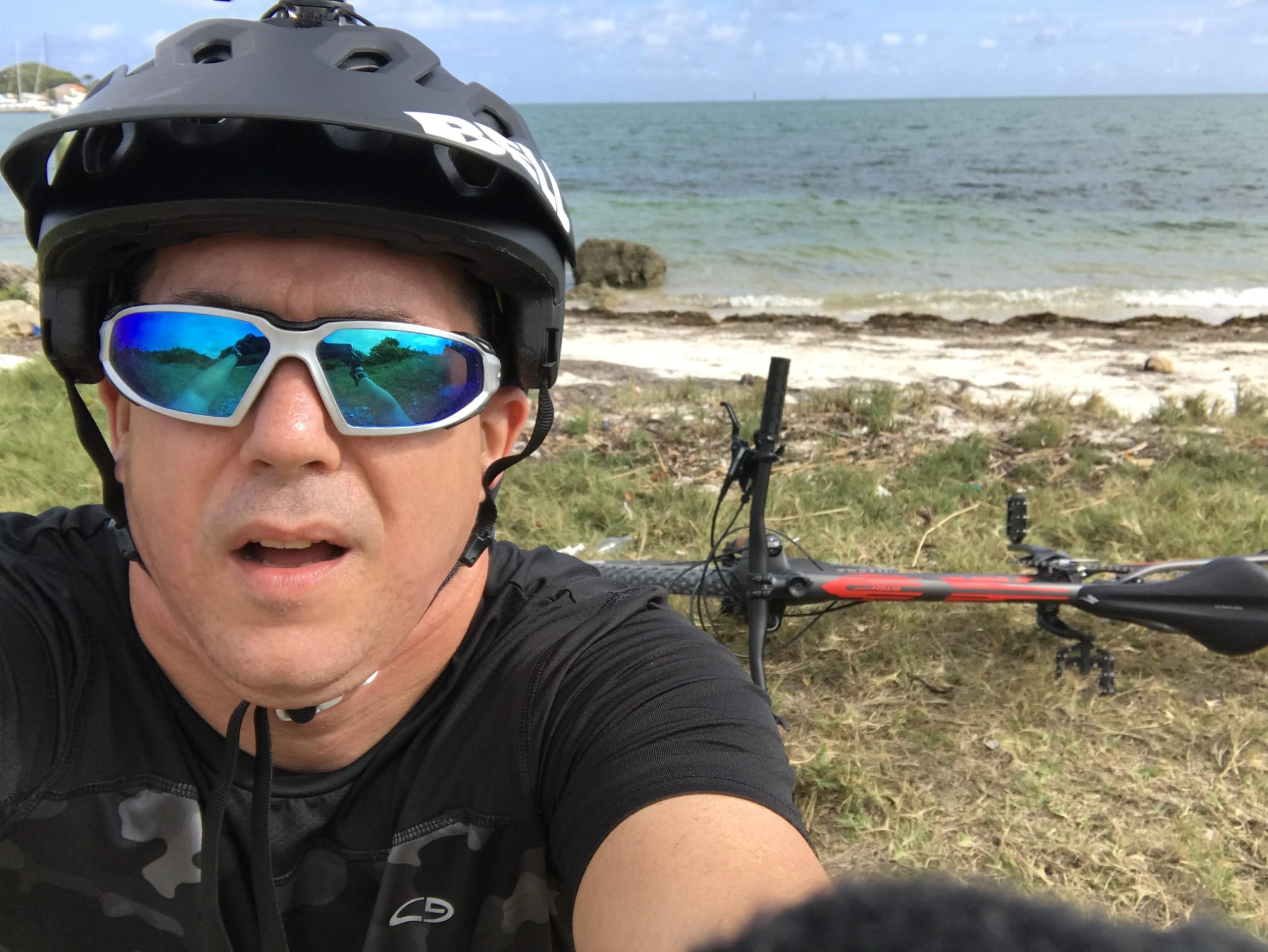 A person wearing a black helmet and reflective sunglasses takes a selfie near the beach, with grassy terrain and water in the background. A black and red bicycle is visible beside them. The sky is partly cloudy and the scene suggests an outdoor recreational activity. Virginia Key North Point mountain bike trail.