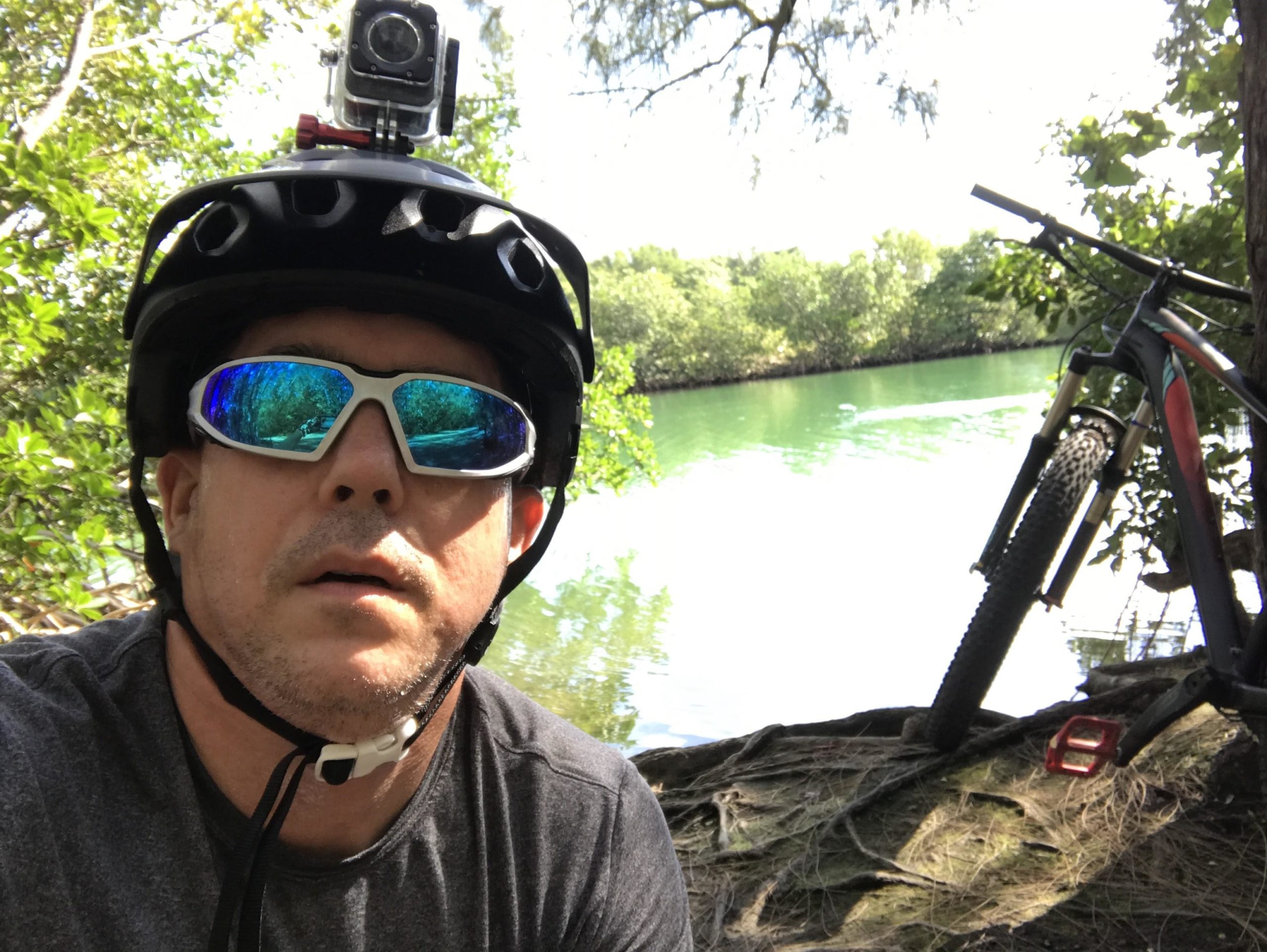 A person wearing a helmet and sunglasses, with a camera mounted on the helmet, poses for a selfie by a tranquil green body of water. A mountain bike is resting nearby, surrounded by lush greenery and trees. Oleta River State Park mountain bike trail.