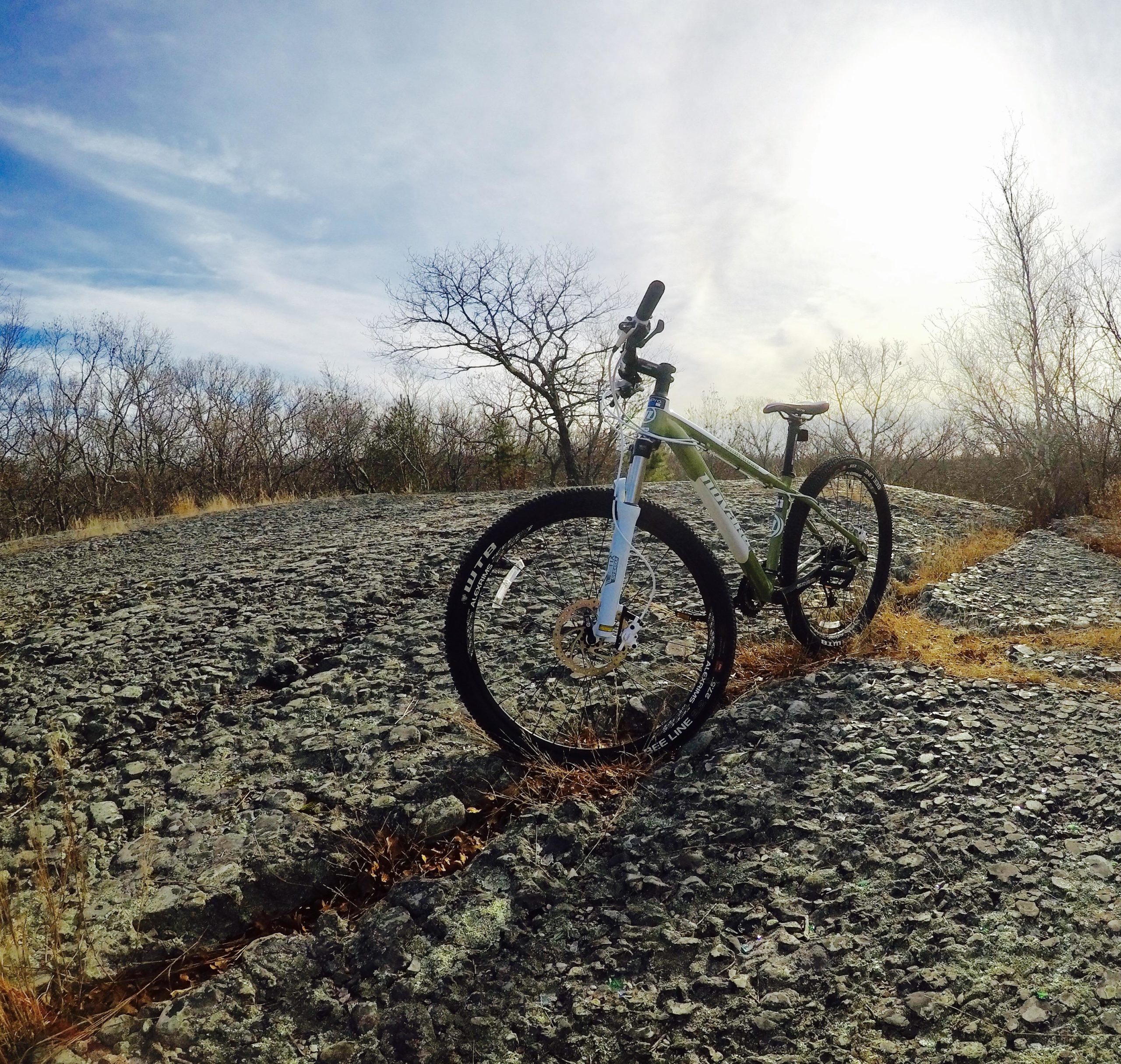 A mountain bike positioned on a rocky surface, surrounded by sparse trees and dry grass, under a bright sky. The sun is shining in the background, casting light on the bike and the terrain. Village Park/ Abrams Rock mountain bike trail.