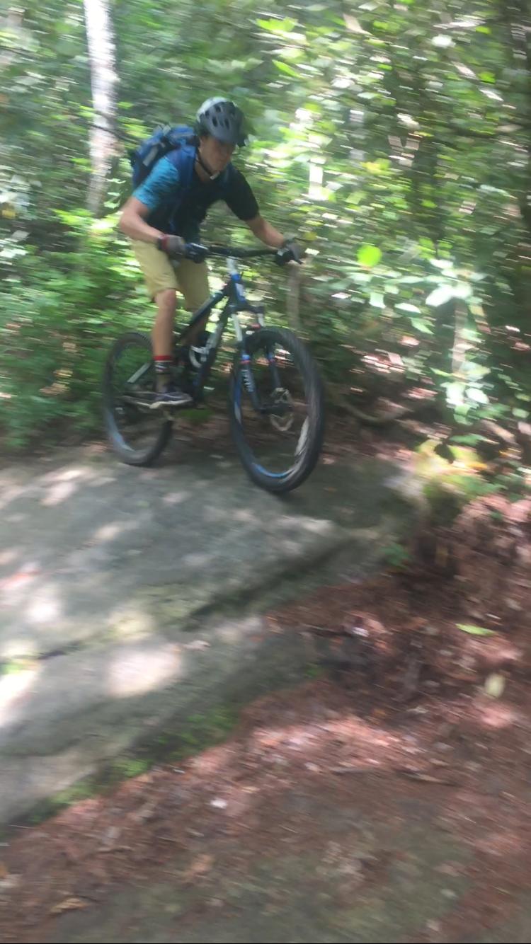 A mountain biker in a helmet and backpack is captured mid-air, jumping off a rocky surface on a wooded trail. The surroundings are lush with green foliage, indicating a natural outdoor environment. The image is slightly blurred, emphasizing the motion and energy of the biking action. DuPont State Forest mountain bike trail.