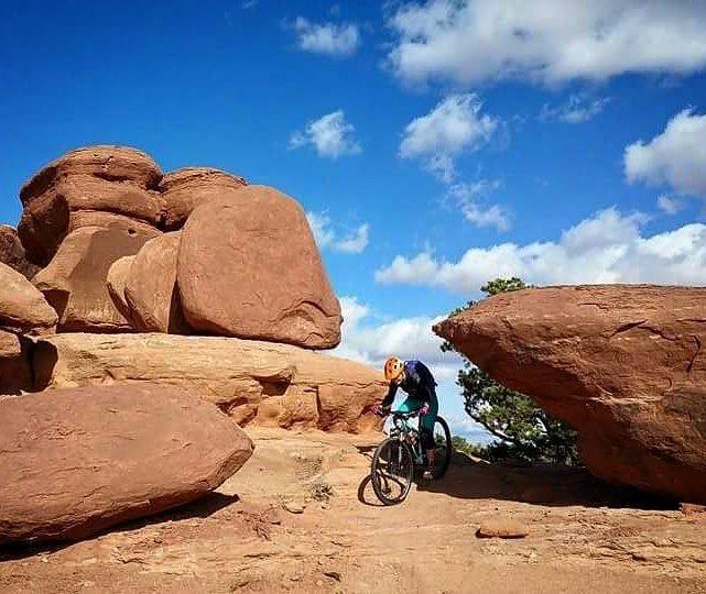 A mountain biker navigating rough terrain surrounded by large red rock formations under a blue sky with scattered clouds. Navajo Rocks mountain bike trail.