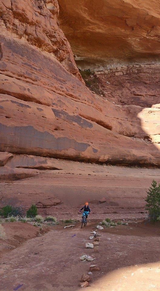 A person riding a mountain bike along a rocky trail beneath a large, red rock formation, surrounded by sparse vegetation and a rugged landscape. Navajo Rocks mountain bike trail.