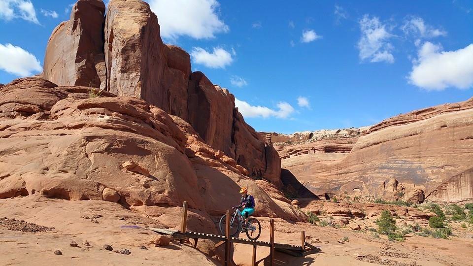 A mountain biker navigating a wooden bridge on a rocky trail, surrounded by towering red rock formations under a bright blue sky with scattered clouds. Navajo Rocks mountain bike trail.