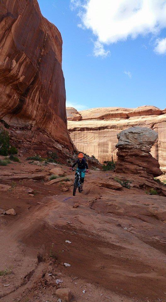 A mountain biker riding along a rocky trail in a desert landscape featuring towering red rock formations and a blue sky with clouds. Navajo Rocks mountain bike trail.