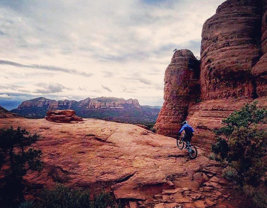 A mountain biker navigating a rocky trail surrounded by large red rock formations and distant mountains under a cloudy sky. Broken Arrow Trail / Chicken Point mountain bike trail.