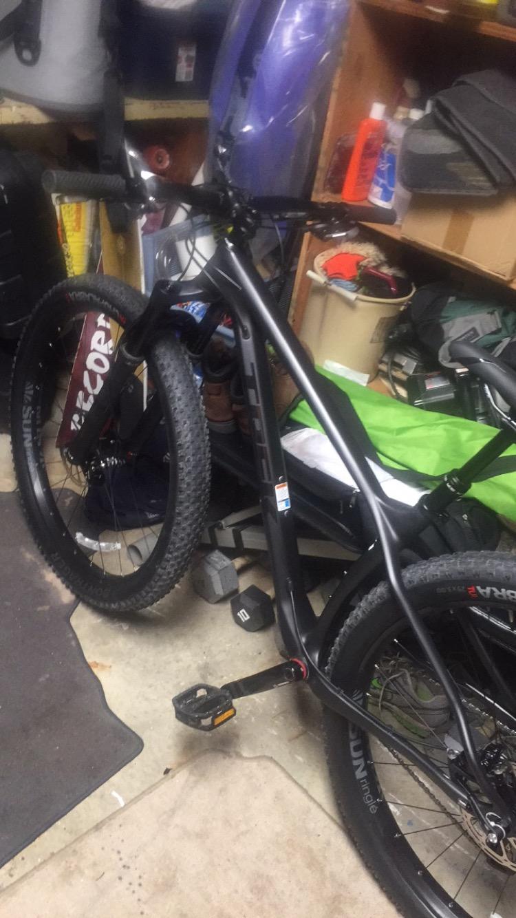 Trek Stache 9 29 Plus: A black mountain bike stands in a cluttered garage, partially obscured by various items. The bike features wide tires and a sturdy frame. In the background, there are containers, tools, and other equipment visible on shelves and the floor.
