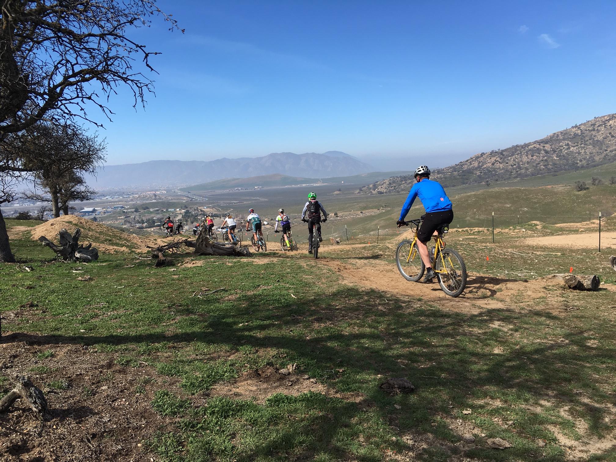 Mountain bikers riding along a dirt trail with a scenic view of rolling hills and a valley in the background on a clear day. The cyclists are mostly dressed in colorful outdoor gear, and a few trees and rocks are visible along the path. TMTA Lehigh trails mountain bike trail.