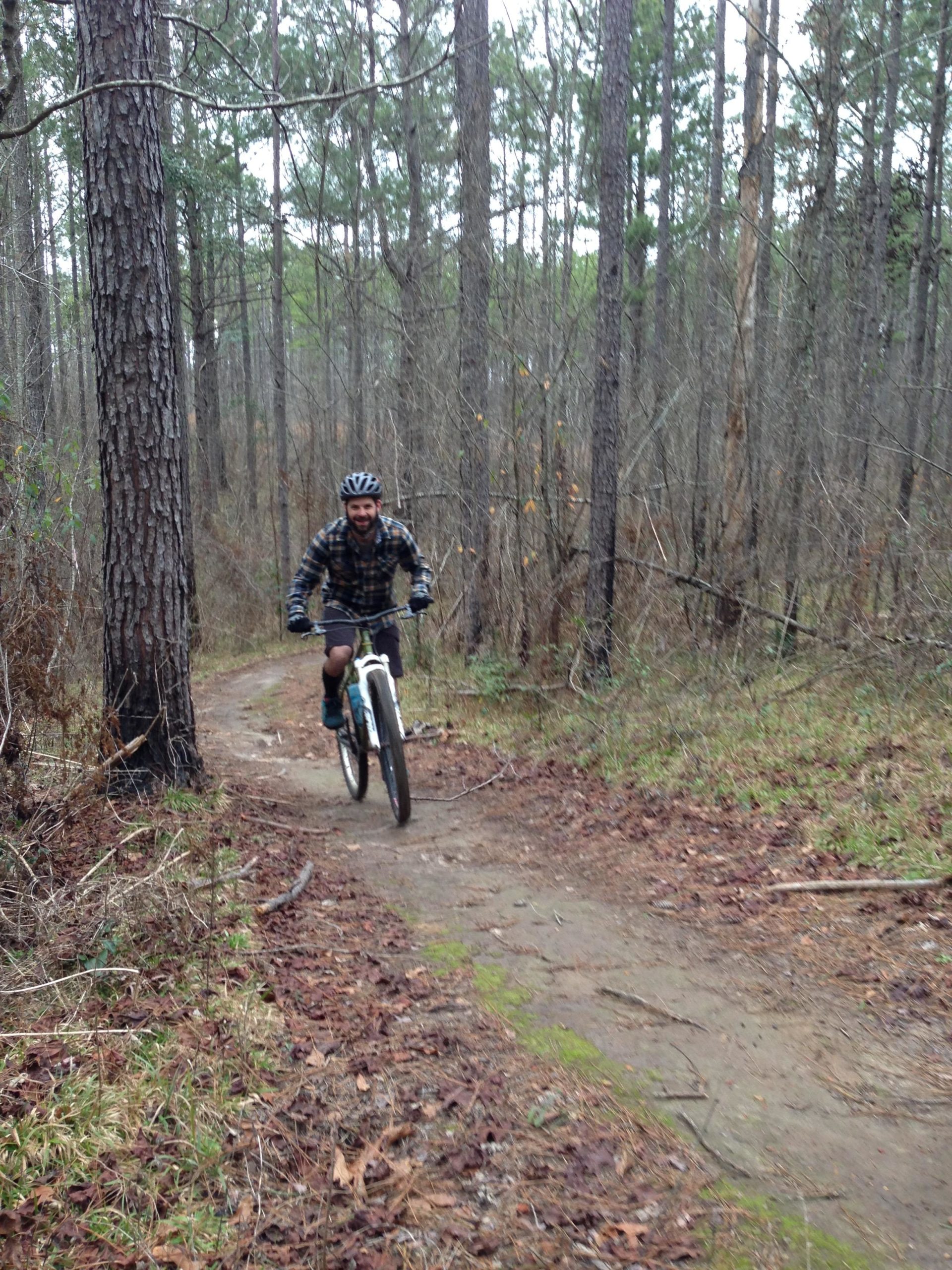 A person riding a mountain bike along a dirt trail in a wooded area, surrounded by tall trees and sparse underbrush, on a cloudy day. The rider is wearing a helmet and a flannel shirt, and appears to be enjoying the ride. Mt. Zion Bike Trails mountain bike trail.