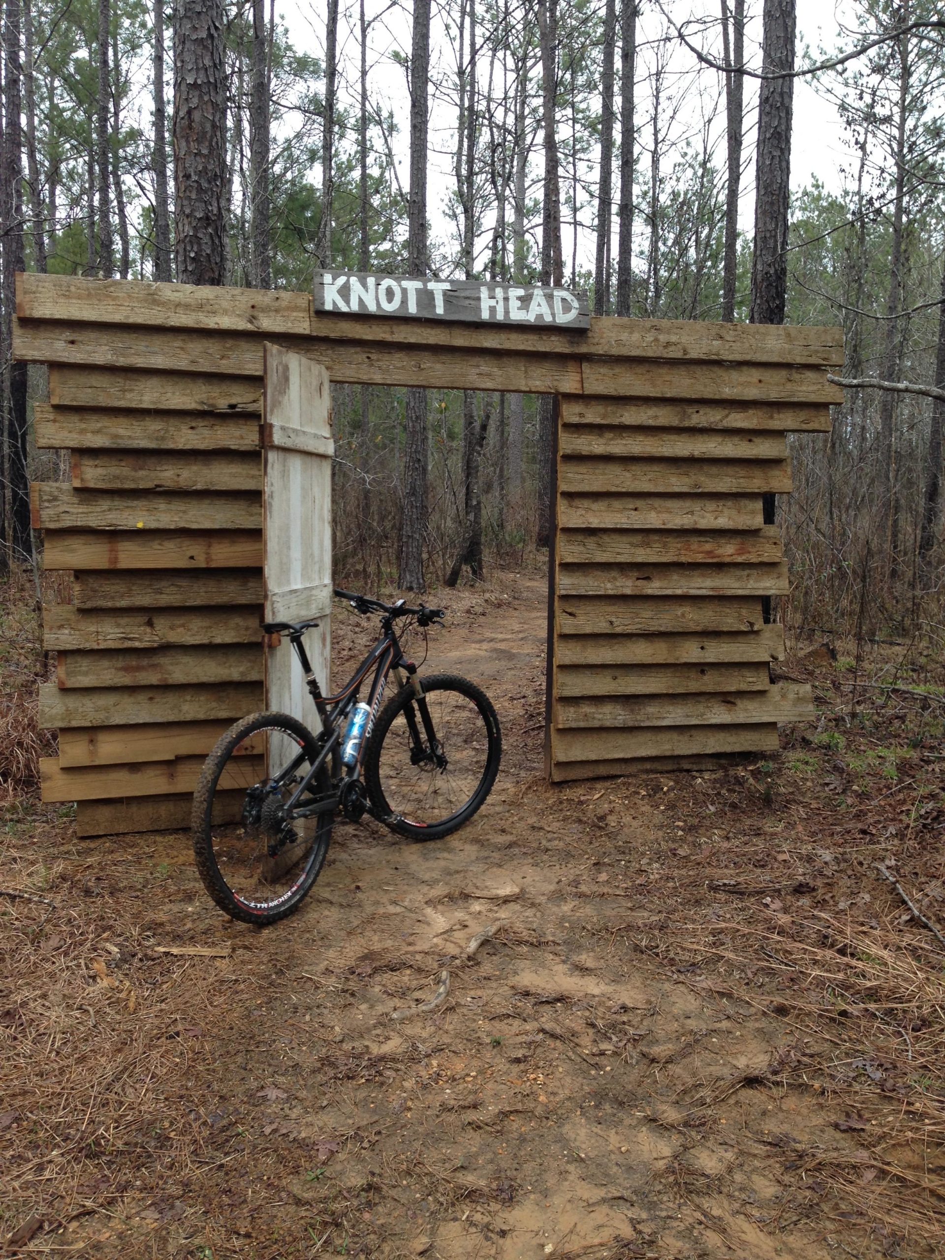 A wooden archway with the sign "KNOTT HEAD" at the top, standing at the entrance to a dirt trail in a pine forest. A mountain bike leans against the structure, with dirt and pine needles on the ground surrounding it. The trees in the background are tall and sparse, creating a natural setting. Mt. Zion Bike Trails mountain bike trail.