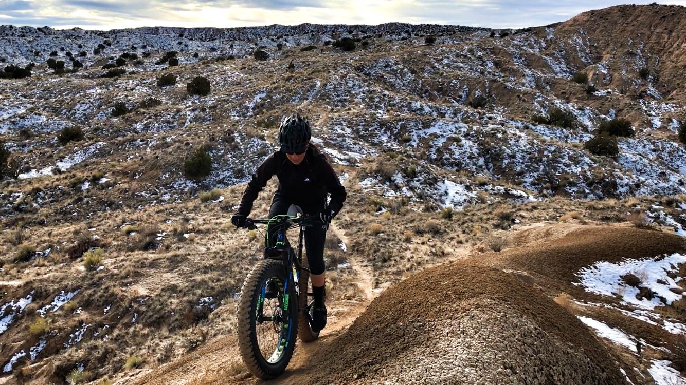 A mountain biker riding a fat bike on a rugged terrain covered with sparse vegetation and patches of snow, ascending a dirt incline against a backdrop of rocky hills under a cloudy sky. Mariposa Fat Bike Trails mountain bike trail.