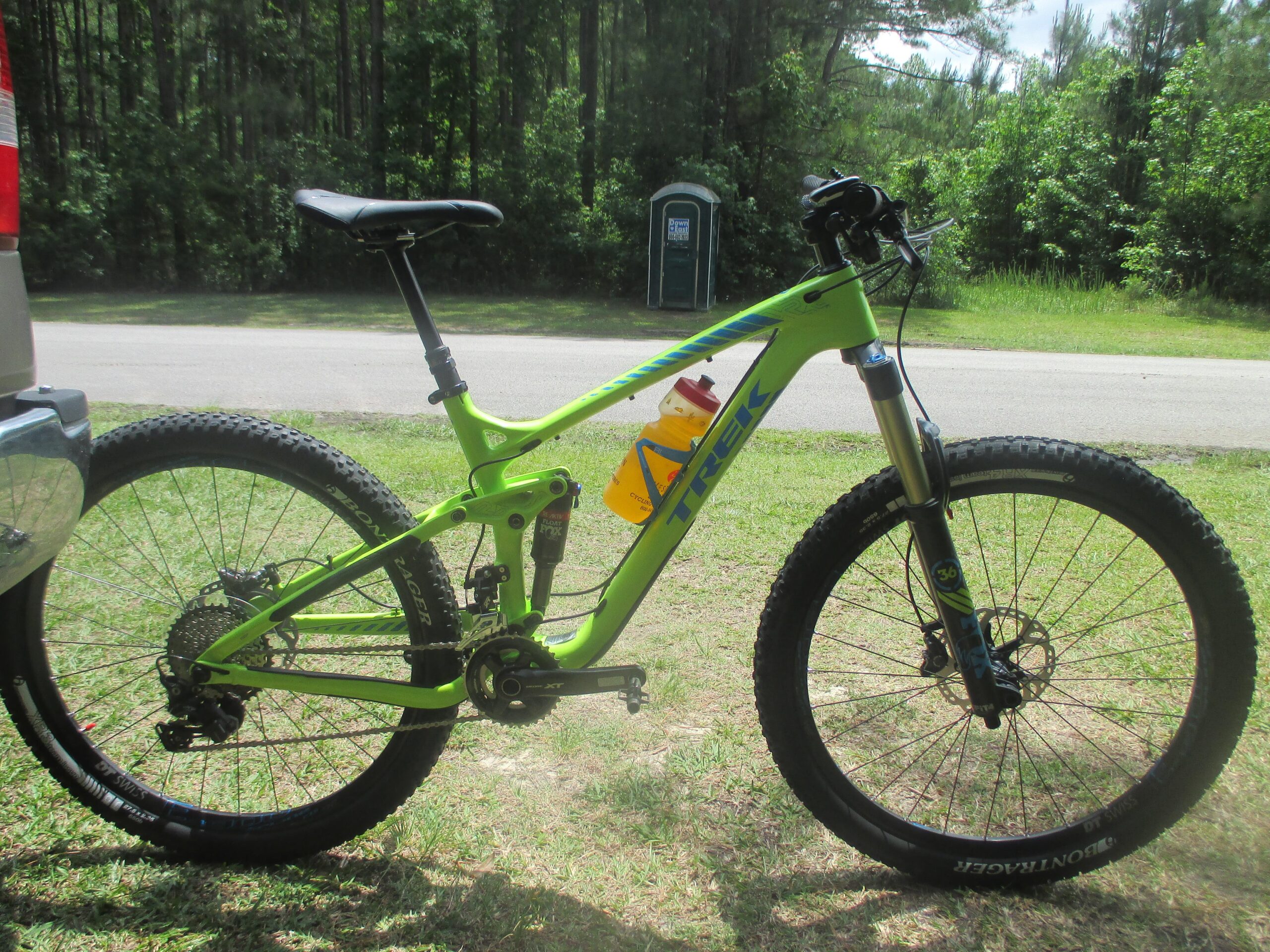 Trek Remedy 9.8 27.5/650b: A bright green Trek mountain bike parked on grass next to a vehicle. The bike features large, rugged tires, a suspension system, and a water bottle holder. In the background, there is a portable restroom and a dense area of trees.