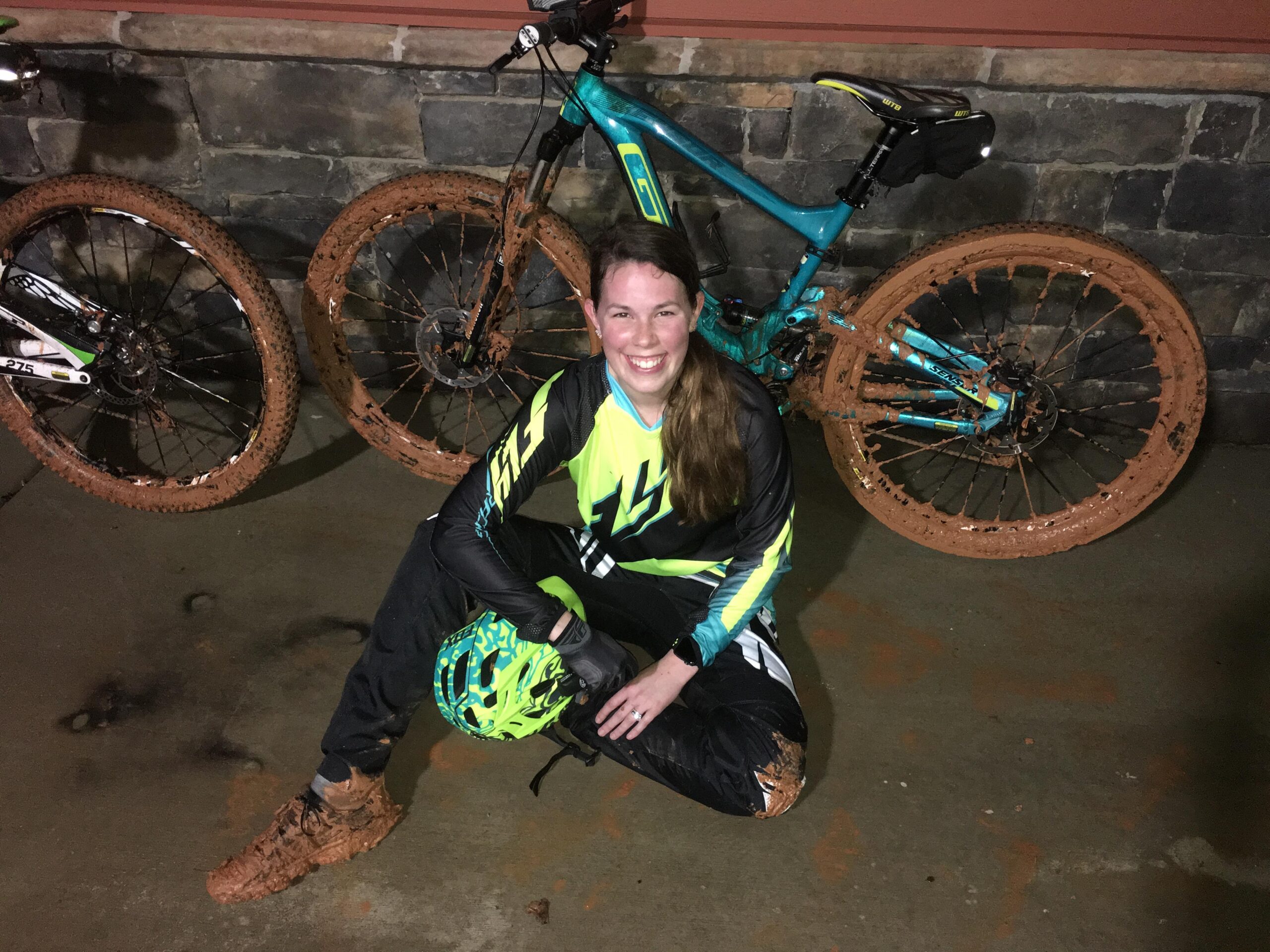 GT Sensor Elite: A woman in a brightly colored cycling outfit sits on the ground with a mud-covered mountain bike behind her. She is wearing a helmet and gloves, and her clothes are also splattered with mud, indicating that she has just finished a muddy bike ride. Two mountain bikes are visible, featuring muddy tires, against a textured stone wall. The scene conveys a sense of adventure and outdoor activity.