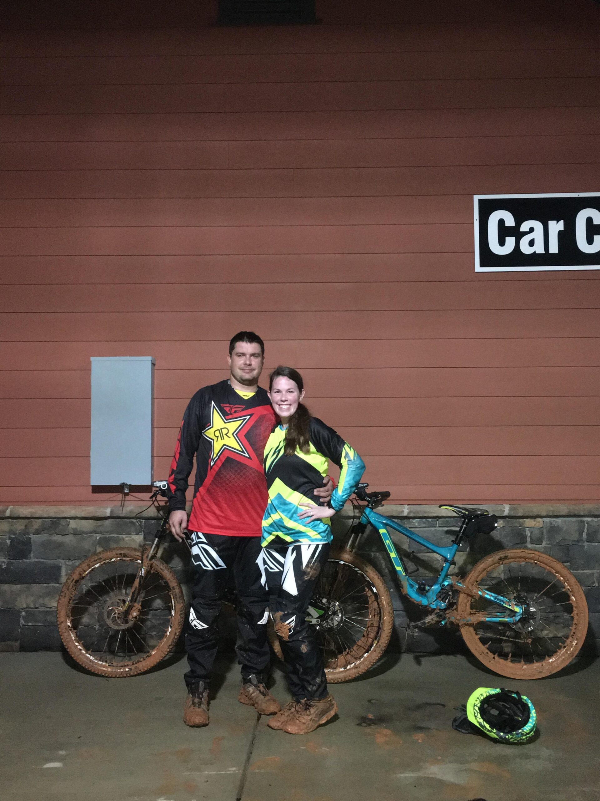 GT Sensor Elite: A couple poses happily together in muddy biking gear, standing beside their mountain bikes. The background features a wooden wall with a sign reading "Car Care." The couple is wearing matching black and colorful outfits, with mud splatters on their clothing and footwear, indicating they've been riding in challenging conditions.
