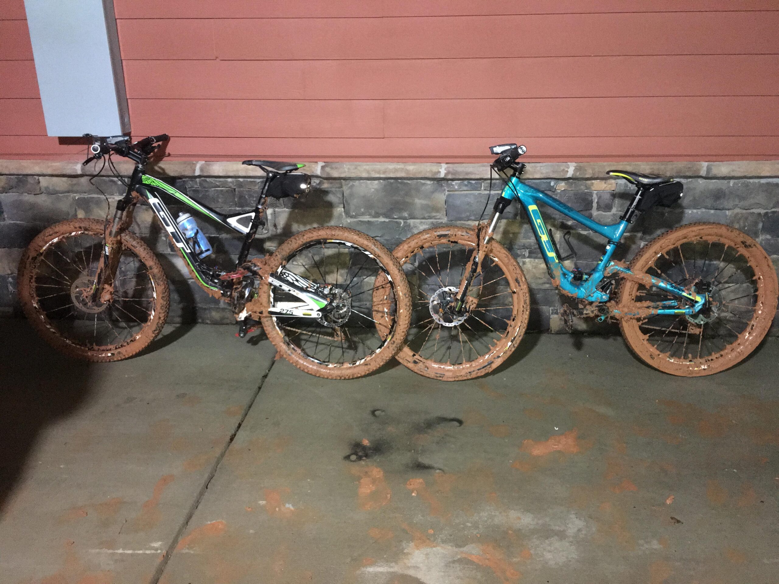 GT Sensor Elite: Two mountain bikes side by side, both covered in mud, are positioned against a stone and wooden wall. The bike on the left is black with green and white accents, while the bike on the right is blue with black details. Both bikes have muddy wheels and frames, indicating recent use in off-road conditions. The setting is dimly lit, with a subtle shine reflecting off the wet surfaces.