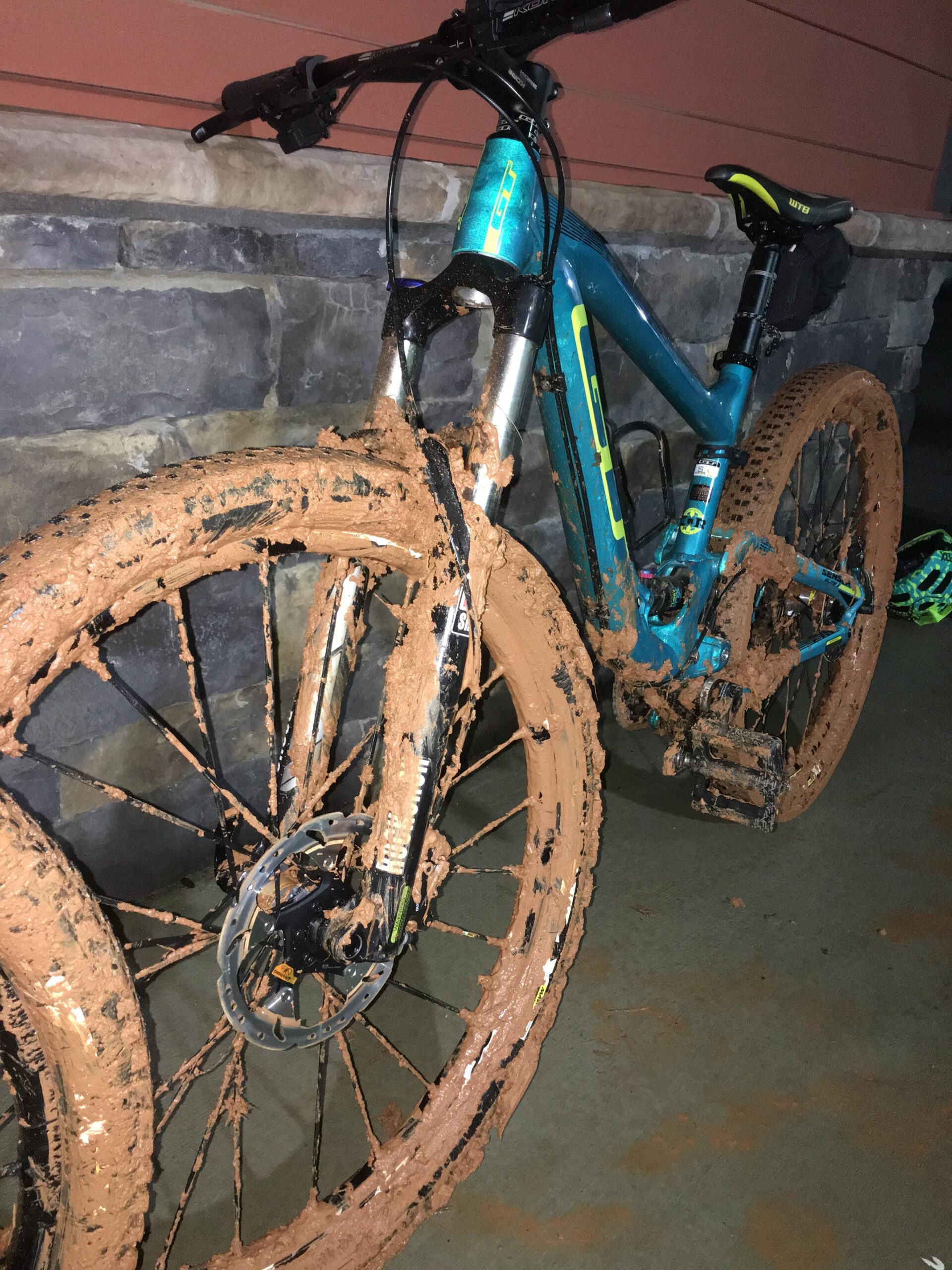 GT Sensor Elite: A mountain bike covered in thick mud, with dirty tires and frame, leaning against a stone wall. The bike has a blue and yellow design, showcasing muddy wheels and components, indicating recent use in muddy terrain.