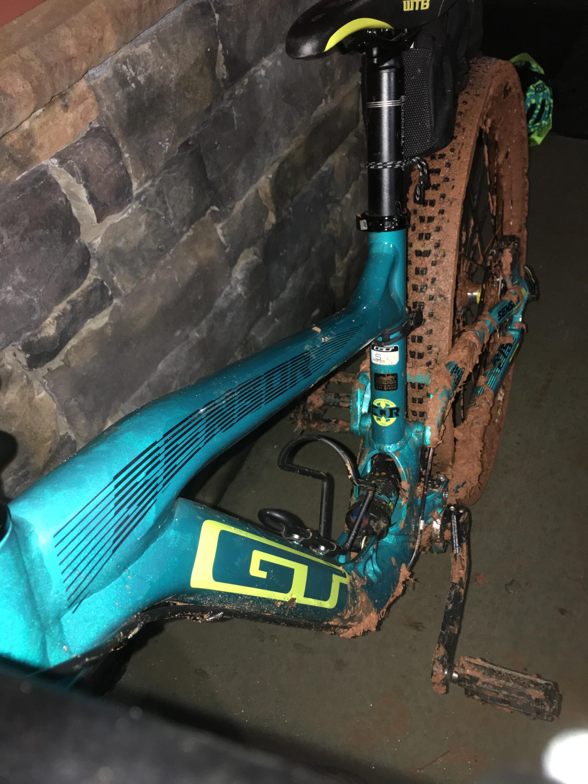 GT Sensor Elite: A close-up view of a teal and yellow GT mountain bike partially covered in mud, leaning against a stone wall. The bike frame is visible alongside the muddy rear wheel and pedal system, showcasing its rugged design suited for off-road cycling.