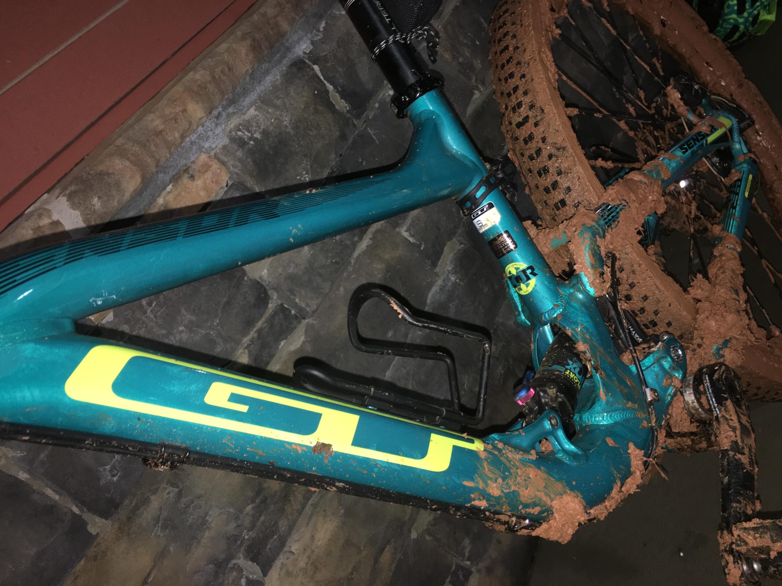 GT Sensor Elite: A close-up view of a mountain bike frame showing a vibrant teal color with yellow accents, covered in thick mud. The bike is positioned on a stone surface, indicating it has just been used in muddy terrain. Parts of the bike, including the wheels and frame, are visibly caked with dirt, highlighting recent outdoor activity.