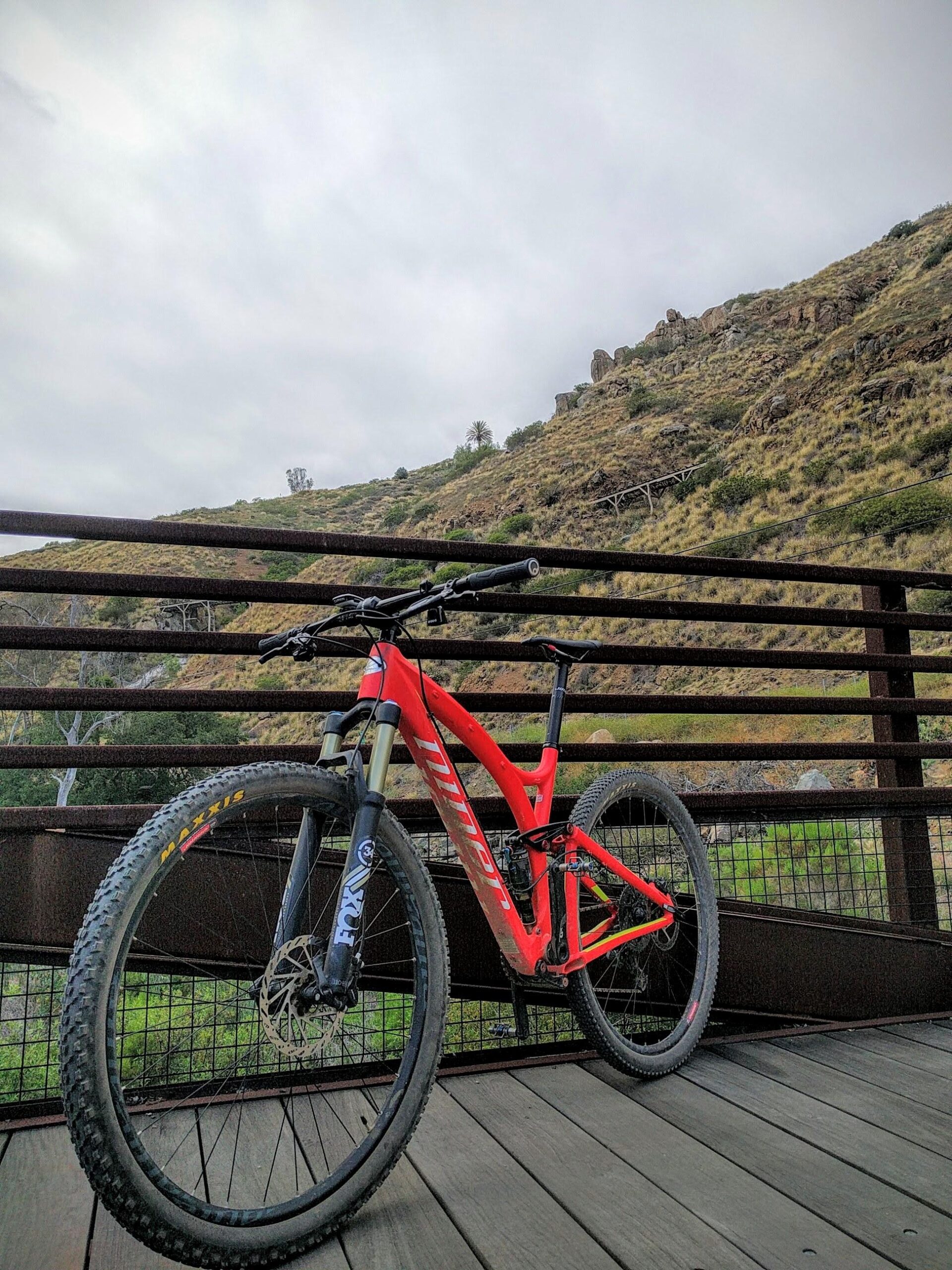 Niner Jet 9 Carbon: A red mountain bike is parked on a wooden bridge with a rugged landscape in the background featuring rolling hills and rocky outcrops under a cloudy sky.