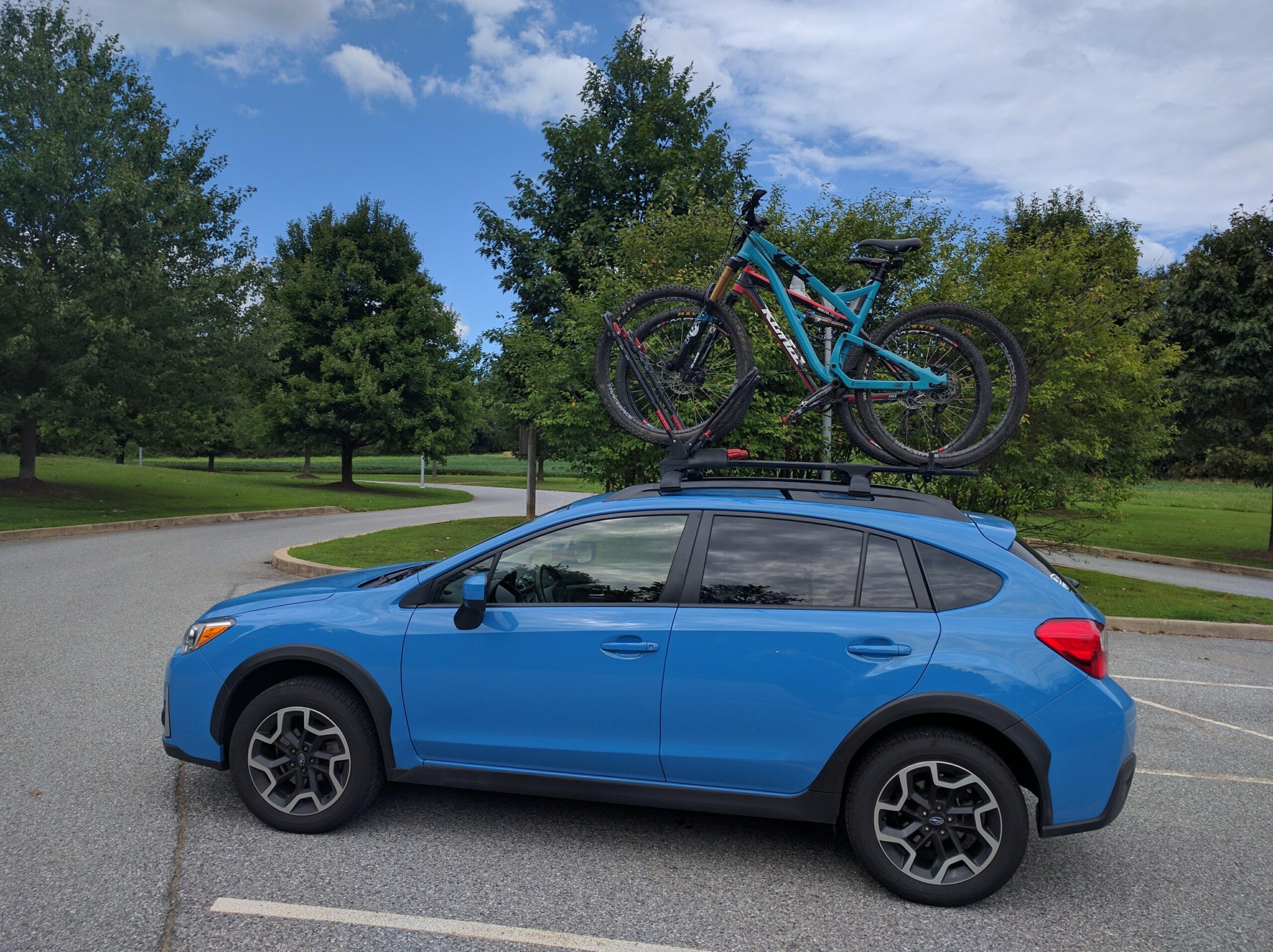 Yeti SB95: A blue SUV parked on a paved area with two mountain bikes secured on the roof rack. The background features green trees and a cloudy sky.