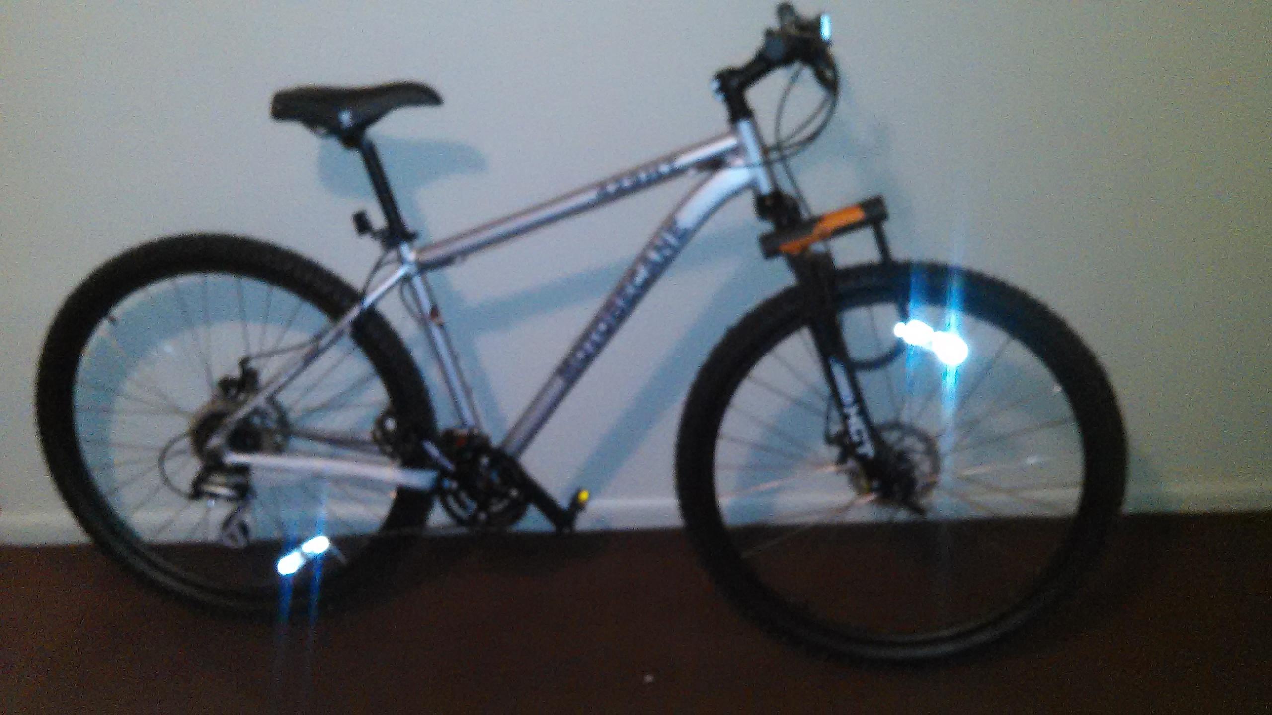 Motobecane 429HT: Mountain bike leaning against a wall, featuring a silver frame, black tires, and reflective details on the wheels. The background is a plain wall with minimal decor.