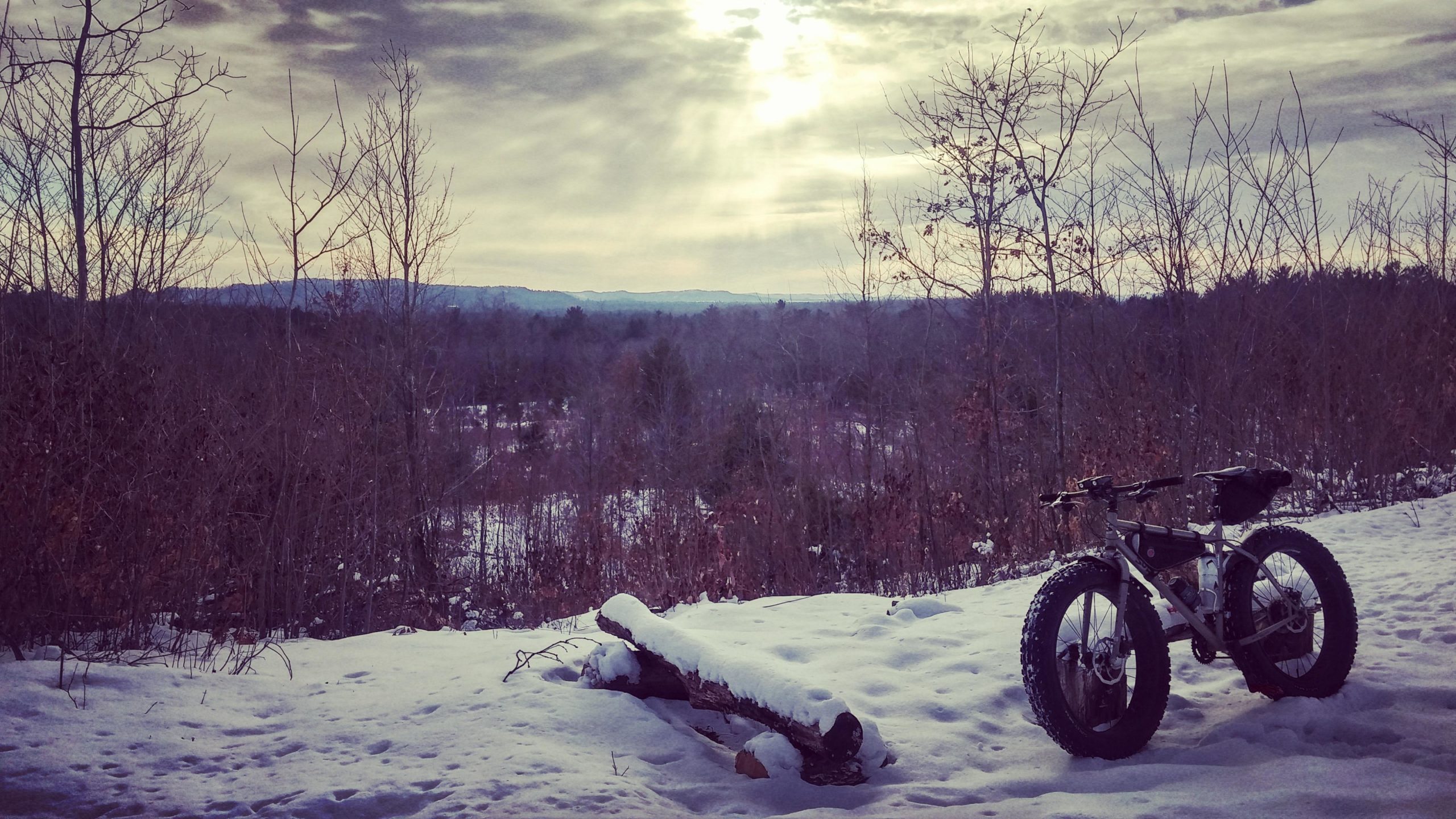 A fat bike resting on a snowy landscape with light snow on the ground, surrounded by bare trees and distant hills under a cloudy sky. The scene captures the tranquility of a winter day. Levis Mounds mountain bike trail.