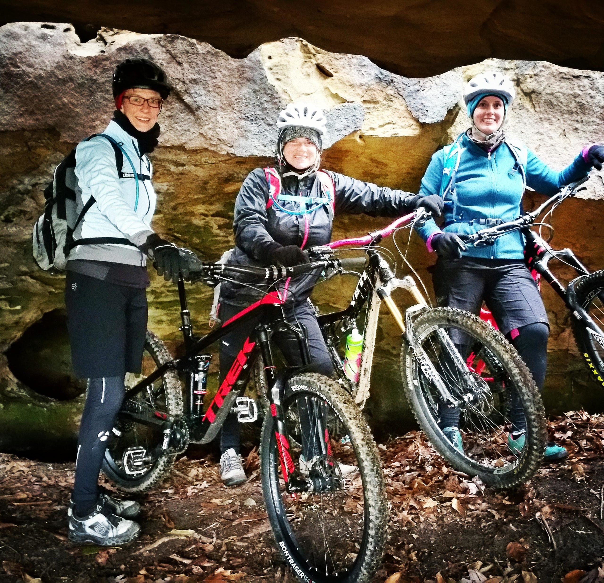Three mountain bikers are posing together under a rocky overhang. They are dressed in outdoor gear, including helmets and gloves, and are standing next to their bikes, which are partially covered in mud. The setting features earthy tones with leaves on the ground and rugged rock formations in the background. Grand Gap mountain bike trail.