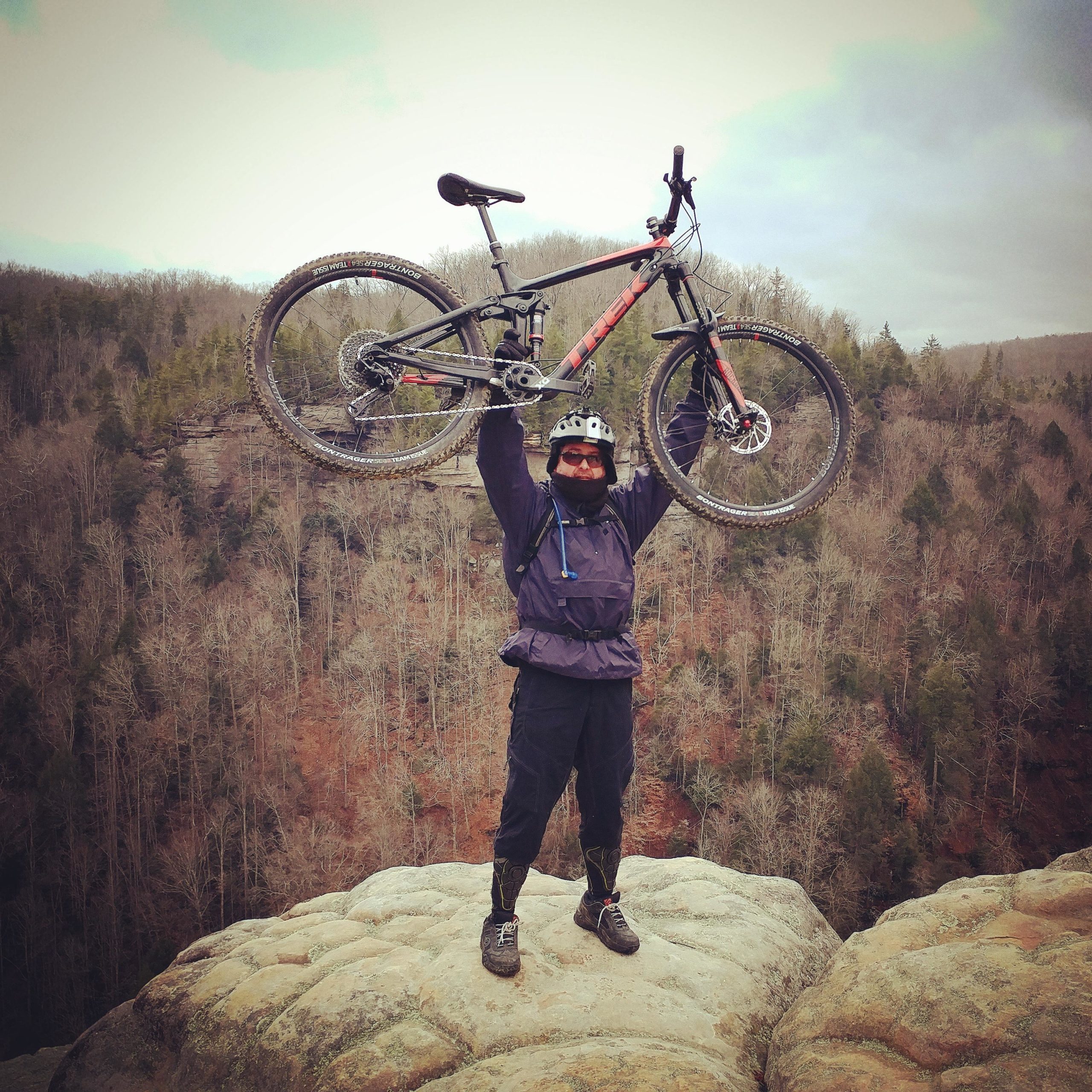 A person wearing a helmet and outdoor gear stands on a rocky ledge, triumphantly holding a mountain bike overhead. Behind them, a wooded landscape stretches out, showcasing a mix of bare trees and greenery against a partly cloudy sky. Grand Gap mountain bike trail.