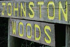 Wooden sign reading "Johnston Woods" in bright yellow letters, mounted on a post surrounded by greenery. Johnston Woods Trails mountain bike trail.