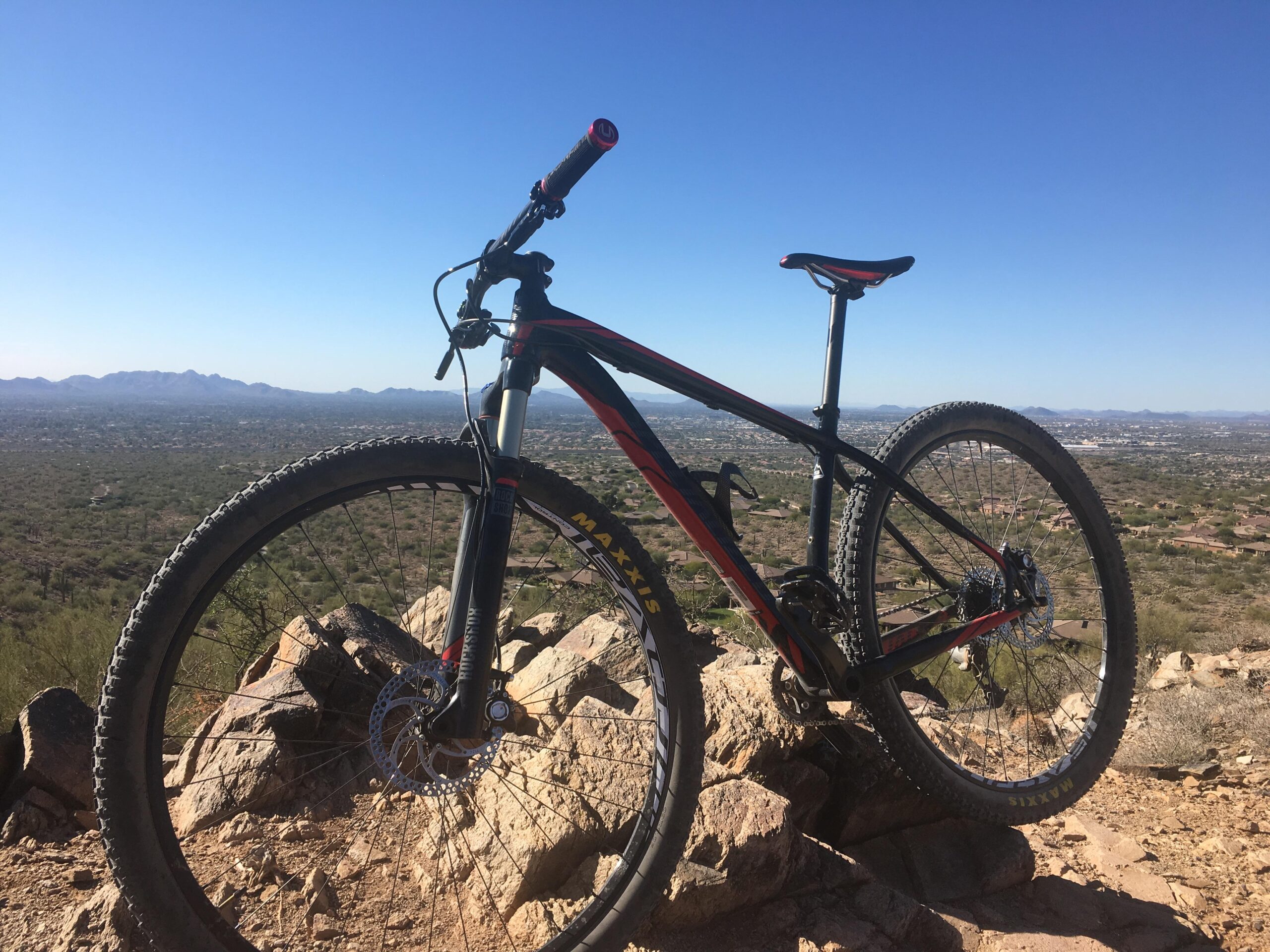 Specialized Stumpjumper FSR Comp 29: A mountain bike propped up on a rocky outcrop, overlooking a desert landscape and distant mountains under a clear blue sky.