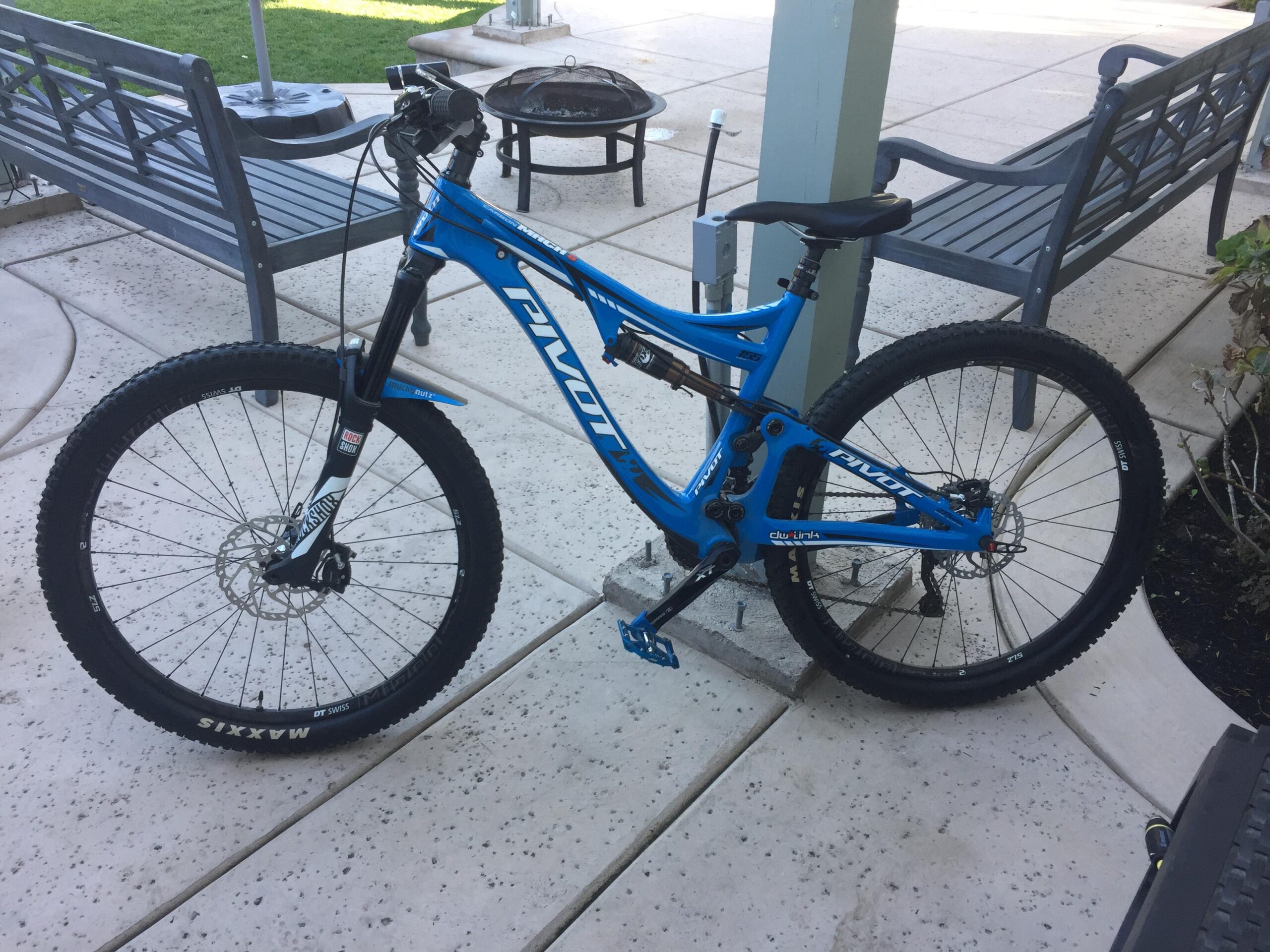 Pivot Mach 6 Carbon: A blue mountain bike with "Pivot" branding stands on a concrete patio. The bike features wide tires and a sturdy frame, alongside a nearby outdoor seating area with wooden benches and a fire pit in the background.