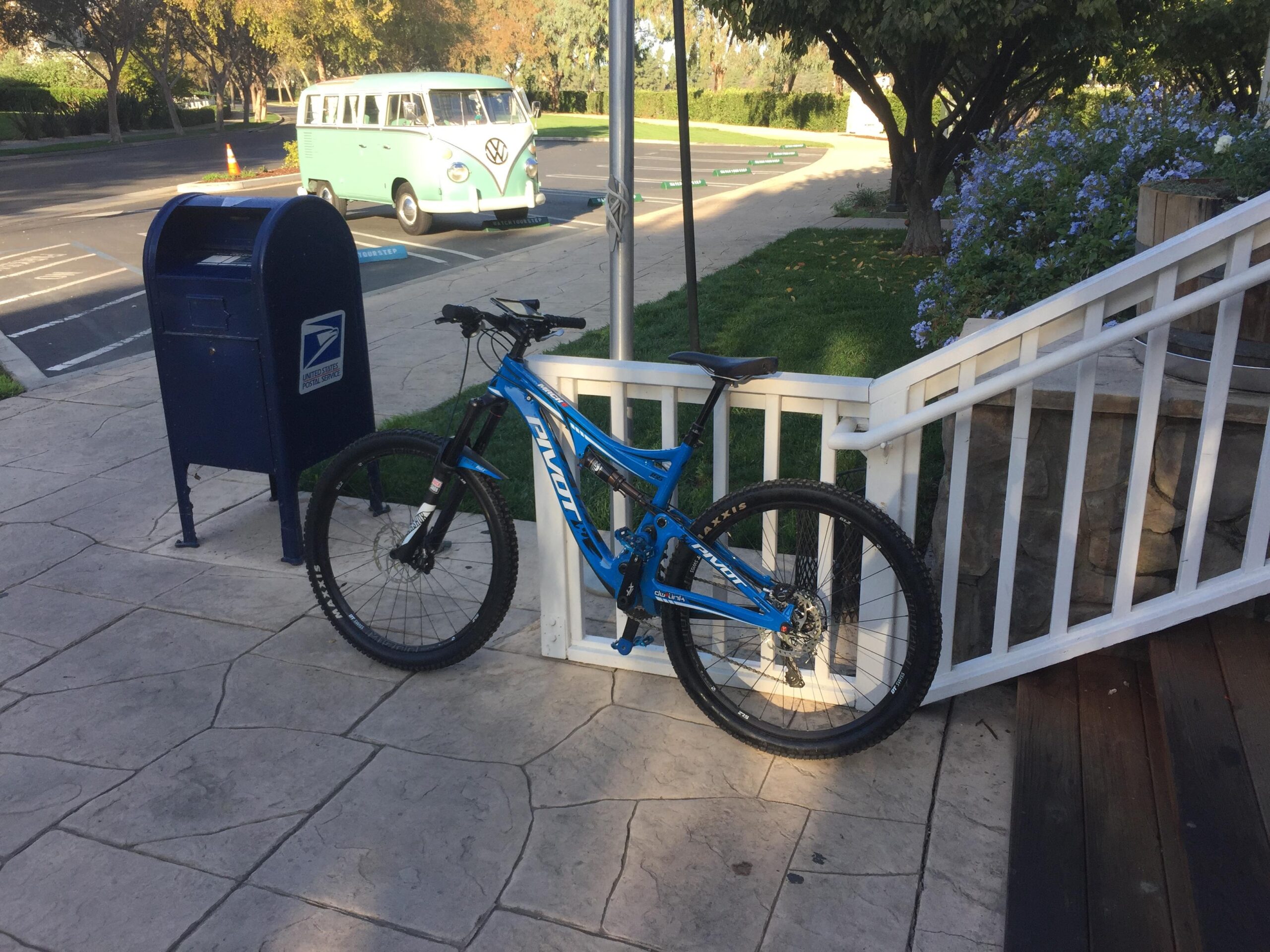 Pivot Mach 6 Carbon: A blue mountain bike is parked next to a blue United States Postal Service mailbox, with a vintage green Volkswagen van visible in the background. The scene is set in a sunny outdoor area with trees and flowers nearby.