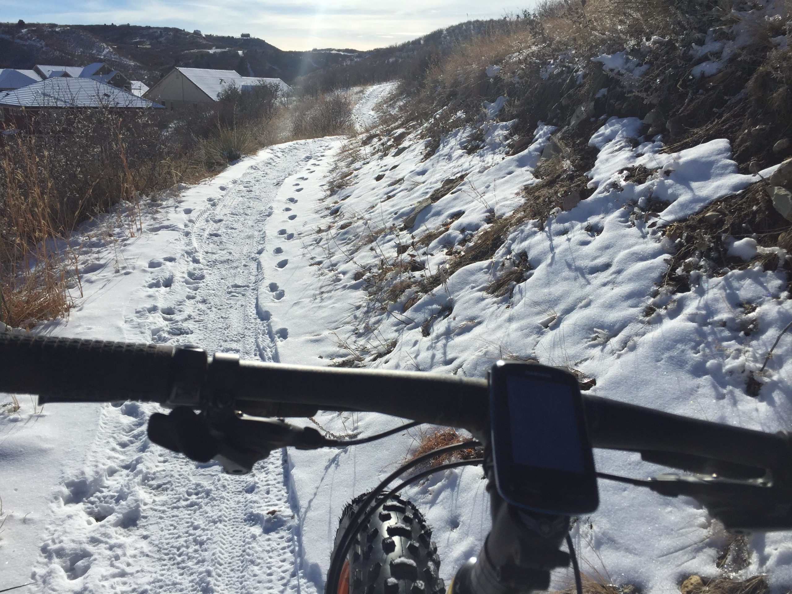 A view from the handlebars of a mountain bike on a snowy trail, with footprints in the snow and houses visible in the background. The handlebars include a bike computer displaying riding data. The scene is set against a backdrop of rolling hills and clear skies. Ridgeline Open Space Trail mountain bike trail.