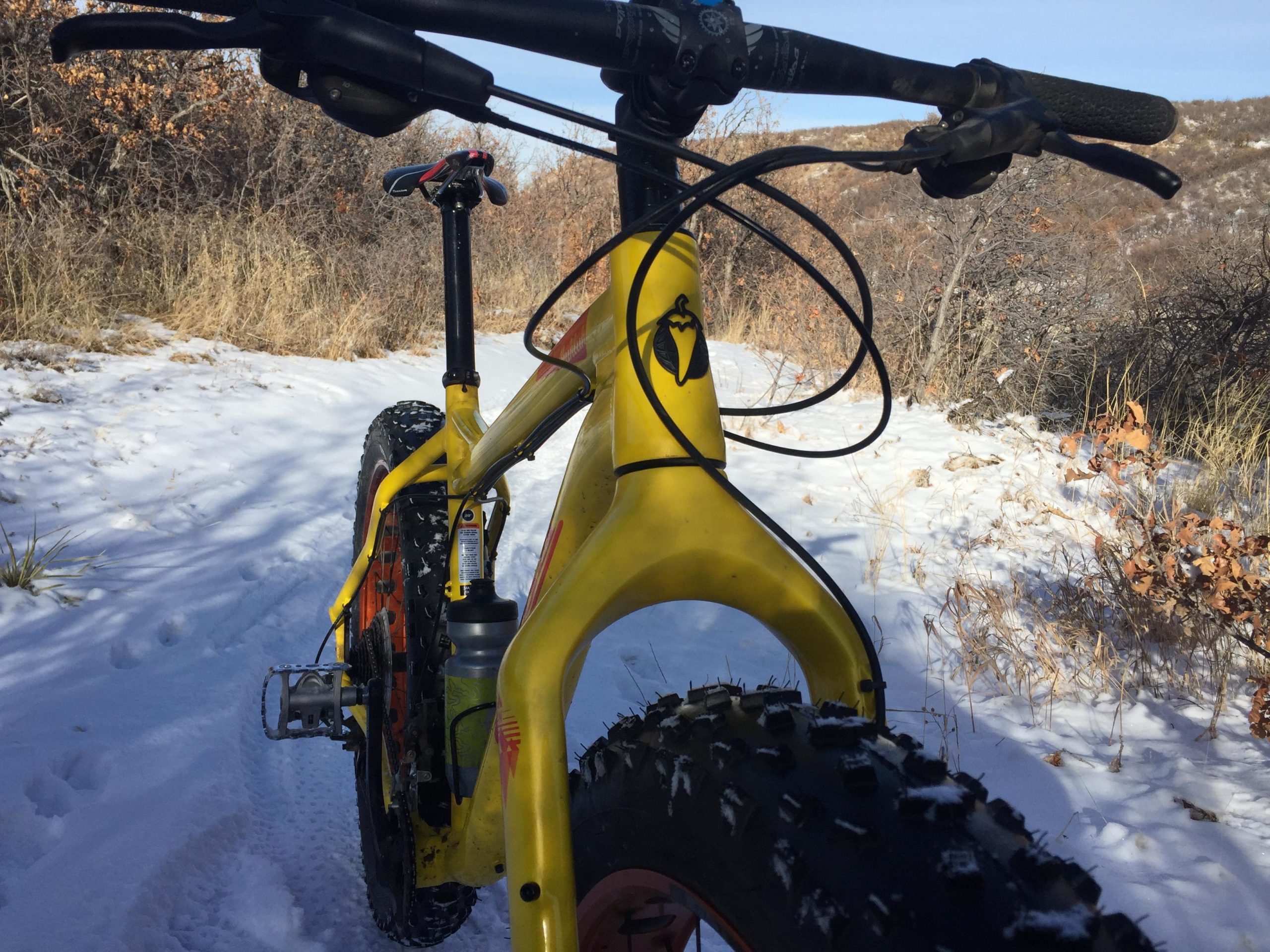 A yellow fat bike with large tires, parked on a snowy trail surrounded by sparse vegetation and dry grasses, with a clear blue sky in the background. Ridgeline Open Space Trail mountain bike trail.