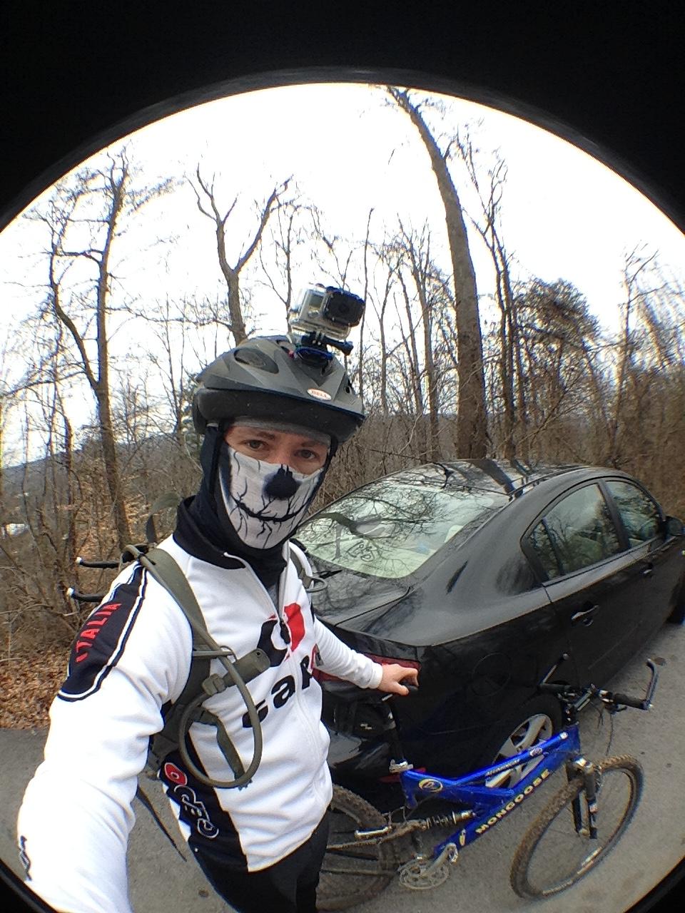 A person wearing a helmet and a face mask stands beside a black car, holding a blue mountain bike. The individual is dressed in a long-sleeve cycling jersey with logos and appears ready for a biking adventure in a wooded area during winter. A camera is mounted on the helmet for recording. Sharps Ridge Memorial Park mountain bike trail.