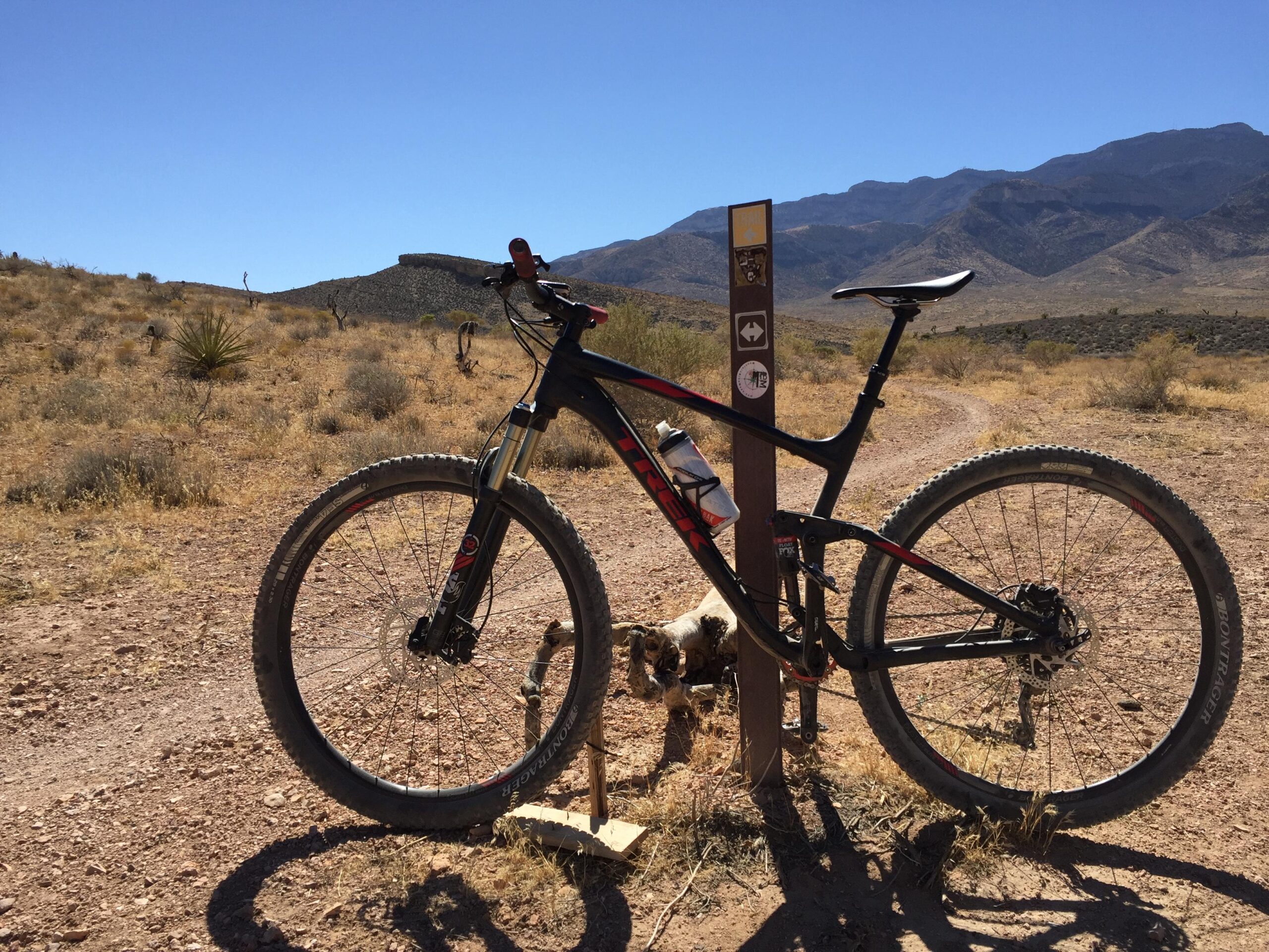 Trek Fuel EX 8: A black mountain bike is leaning against a trail marker on a dirt path, surrounded by dry grass and shrubs. In the background, there are mountains under a clear blue sky, and a small dog is visible near the bike.