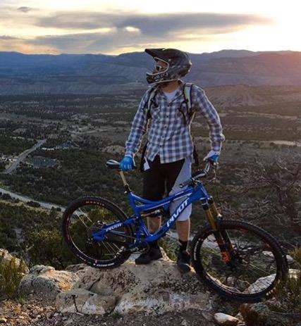 Pivot Mach 6 Carbon: A person in a plaid shirt and protective gear stands next to a blue mountain bike on a rocky outcrop, overlooking a scenic landscape at sunset. The sky is filled with clouds and hints of orange and pink, while the valley below features greenery and winding roads.