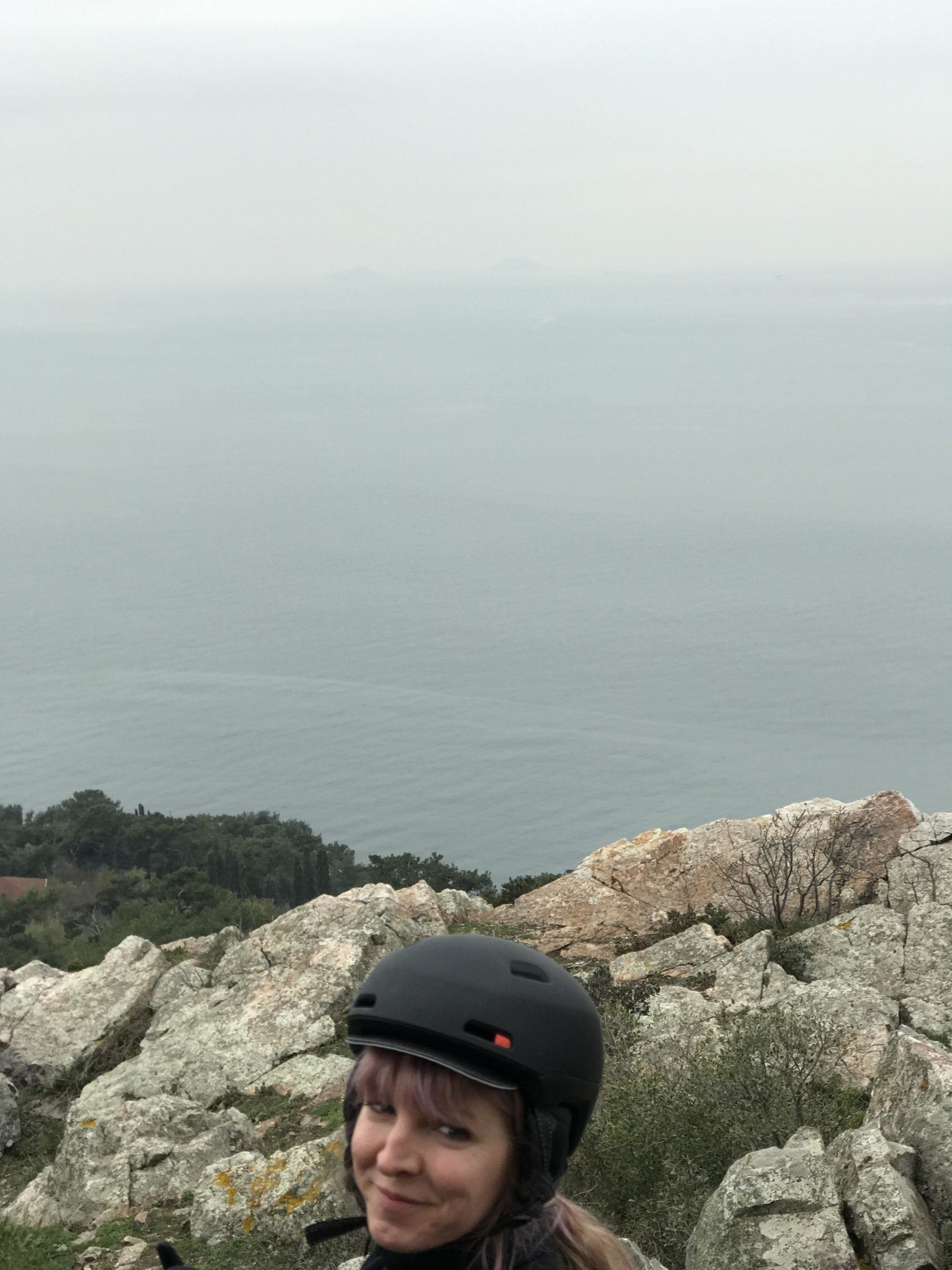 A person with pink hair wearing a black helmet smiles in front of a rocky landscape and a calm sea under a cloudy sky. Distant islands are visible on the horizon. Aya Yorgi Downhill mountain bike trail.