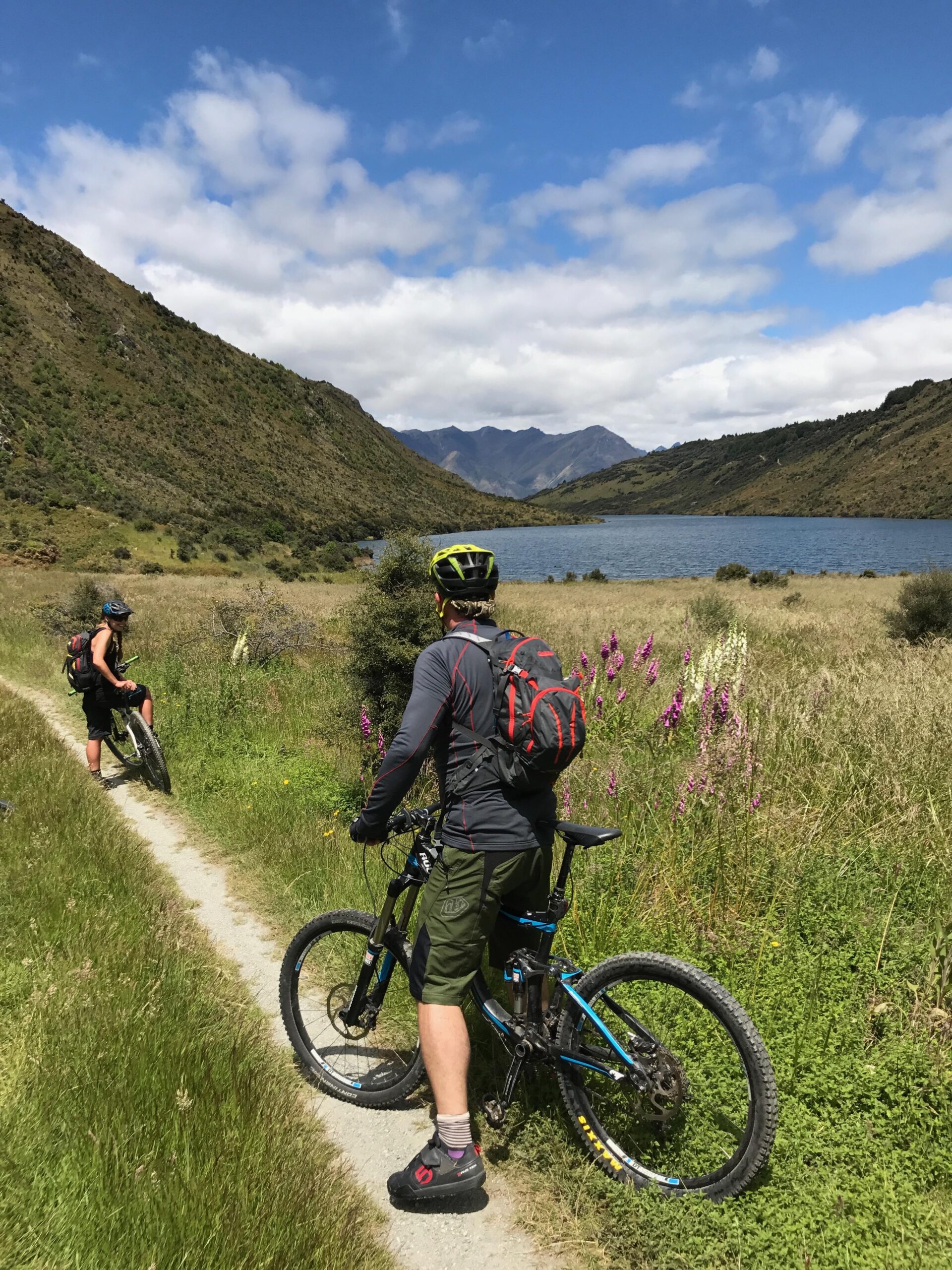 Rocky Mountain Slayer 70: Two mountain bikers pause on a gravel trail surrounded by lush greenery and wildflowers, with a serene lake and rolling hills in the background under a partly cloudy sky.