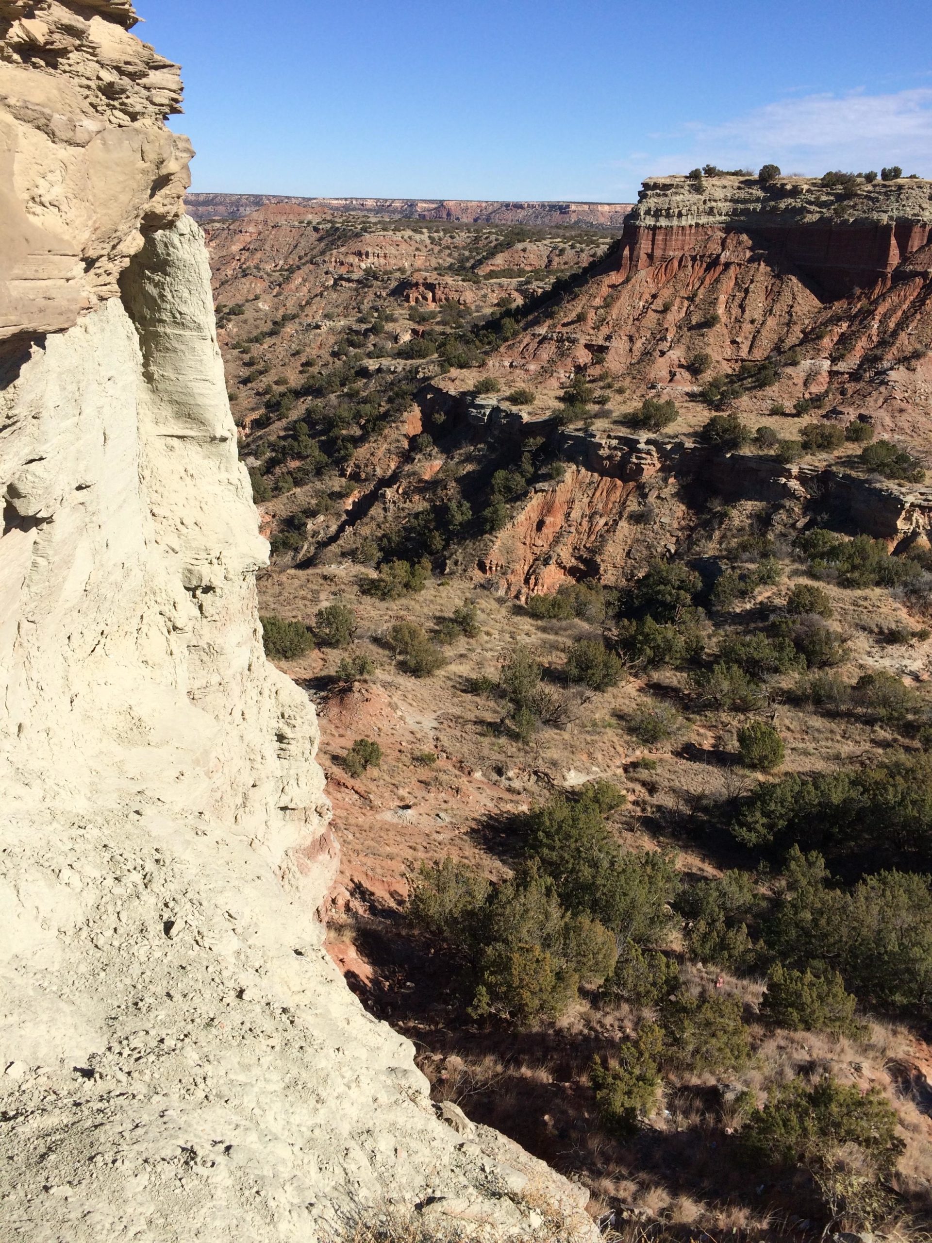 A panoramic view of a rugged canyon landscape, featuring steep rock formations in shades of red and tan, with sparse vegetation scattered throughout the area. The sky is clear with a few clouds, showcasing a bright blue background. The image captures the natural beauty and geological features of the terrain. Palo Duro Canyon mountain bike trail.